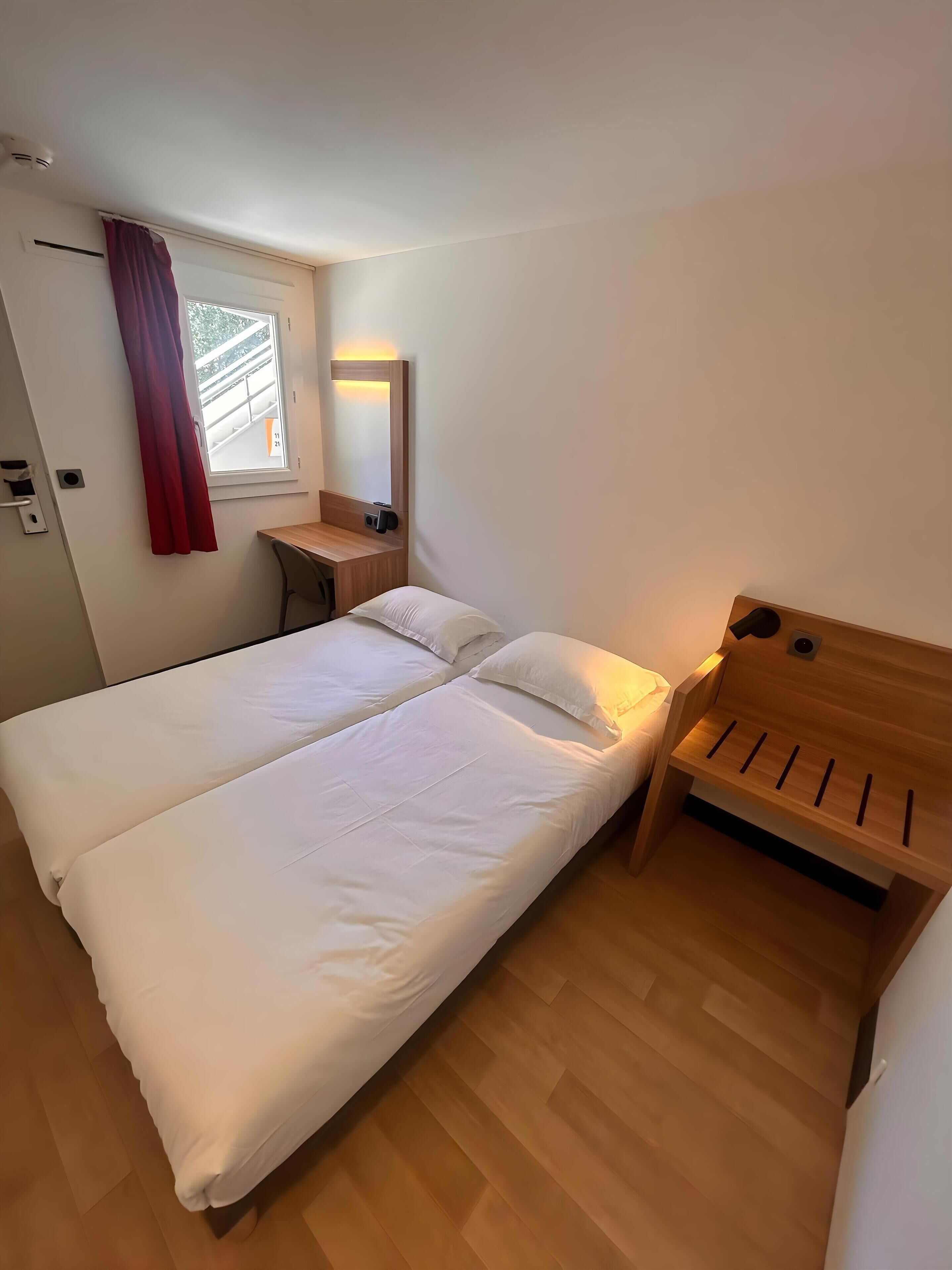A bedroom with a double bed, a wooden bench, a desk, and a window with curtains.