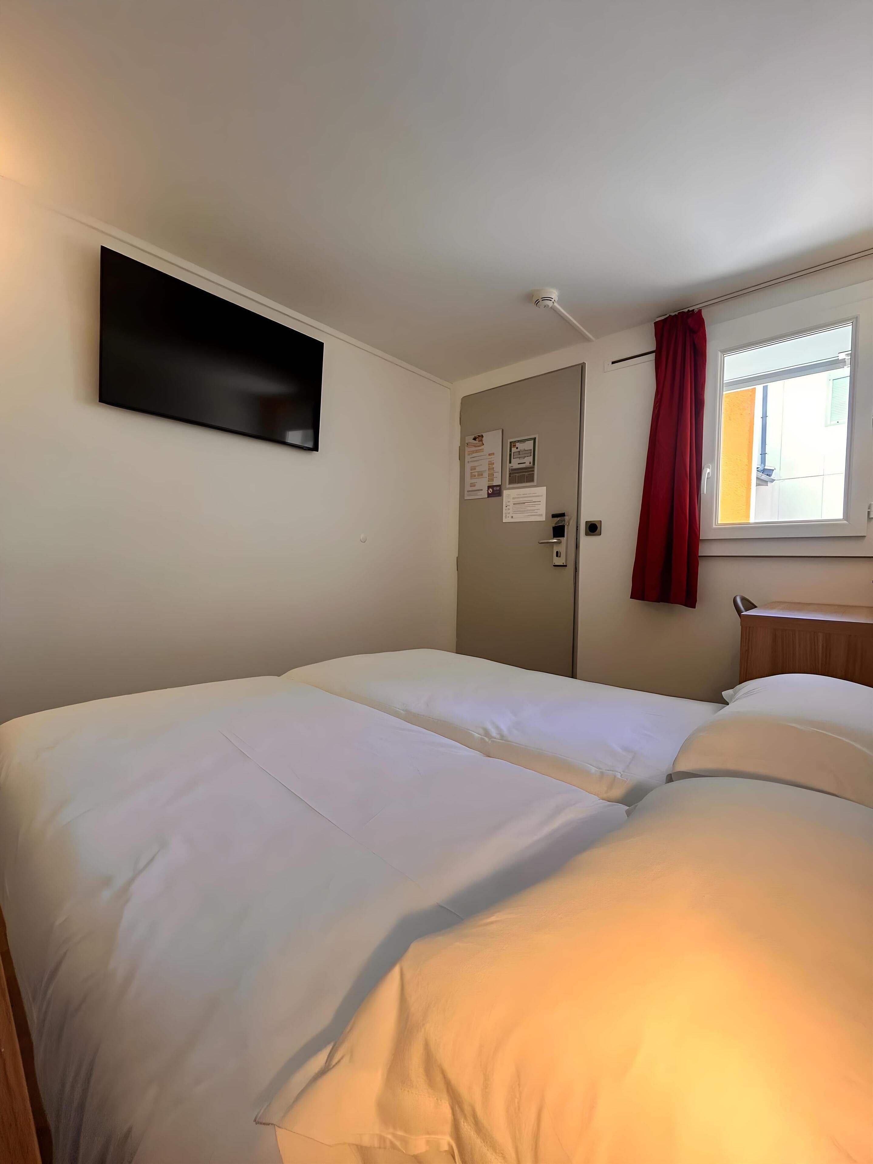 A hotel room with two beds, a flat-screen TV mounted on the wall, a door with a window, and a red curtain.