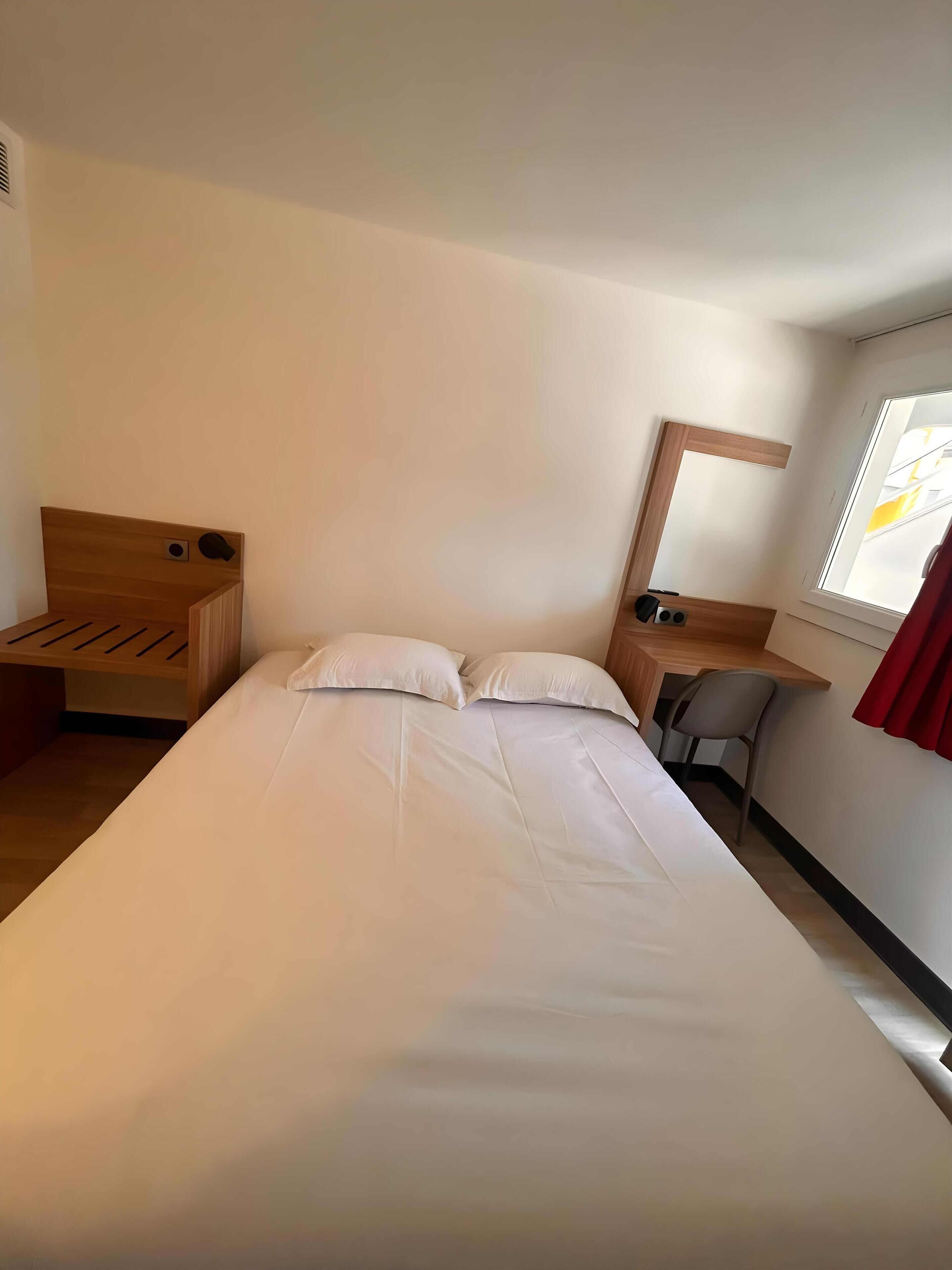 A hotel room with a bed, a desk, a chair, a mirror, and a window.