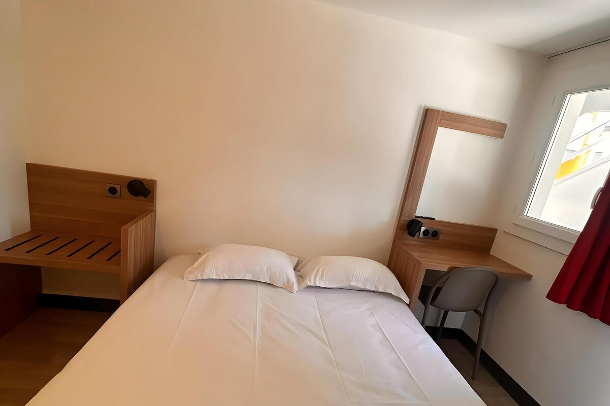 A hotel room with a bed, a desk, a chair, a mirror, and a window.