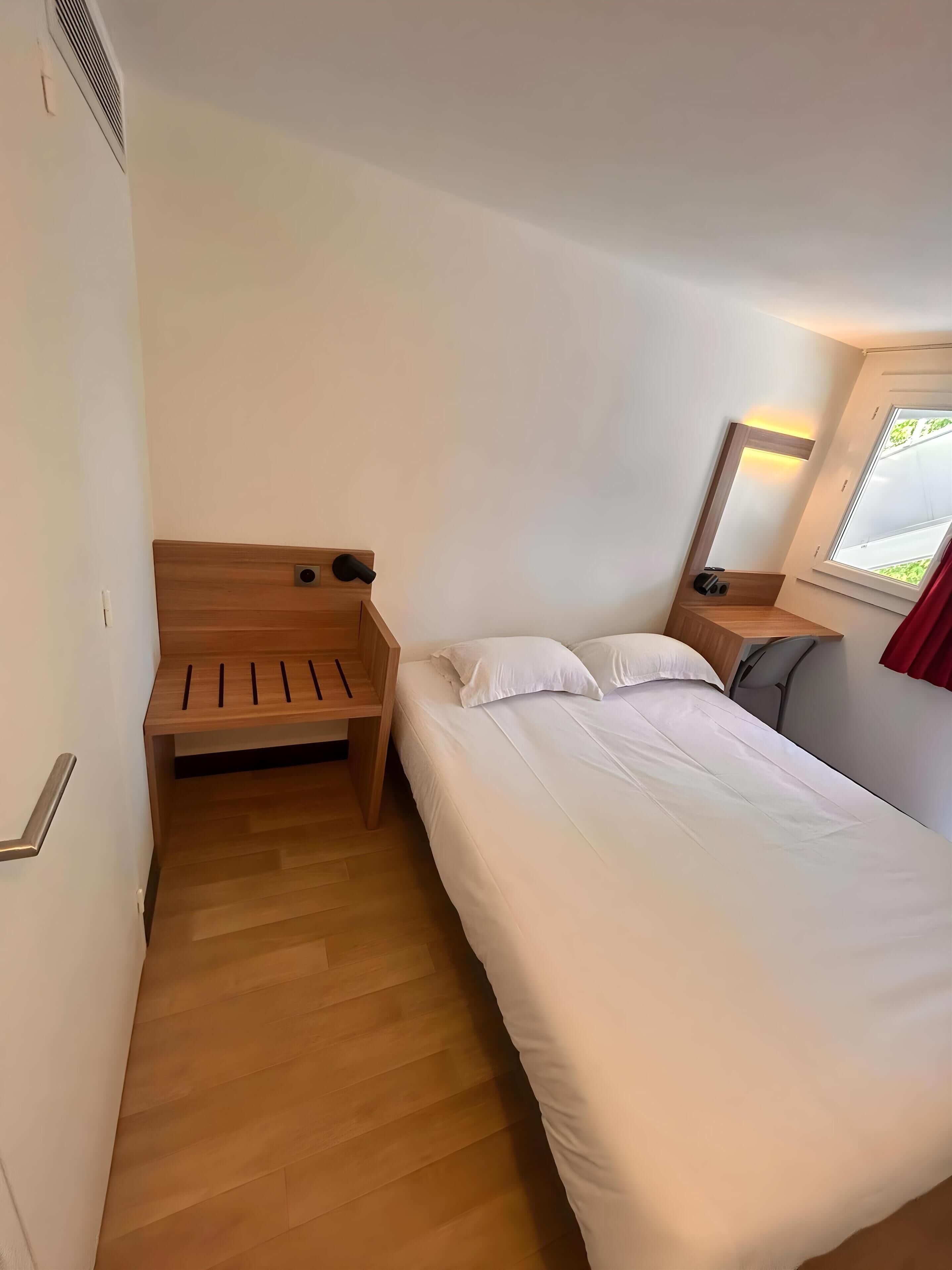 Una habitación de hotel con cama, un banco de madera, una mesita de noche y una ventana con cortinas.