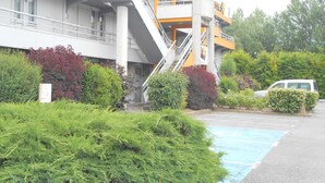 Un edificio de varios pisos con una escalera que conduce a una plataforma amarilla, rodeado de vegetación y un estacionamiento.