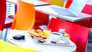 Una bandeja de desayuno con pan, jugo, café y condimentos sobre una mesa en un café de colores vivos, con sillas anaranjadas y una computadora portátil en una mesa cercana.