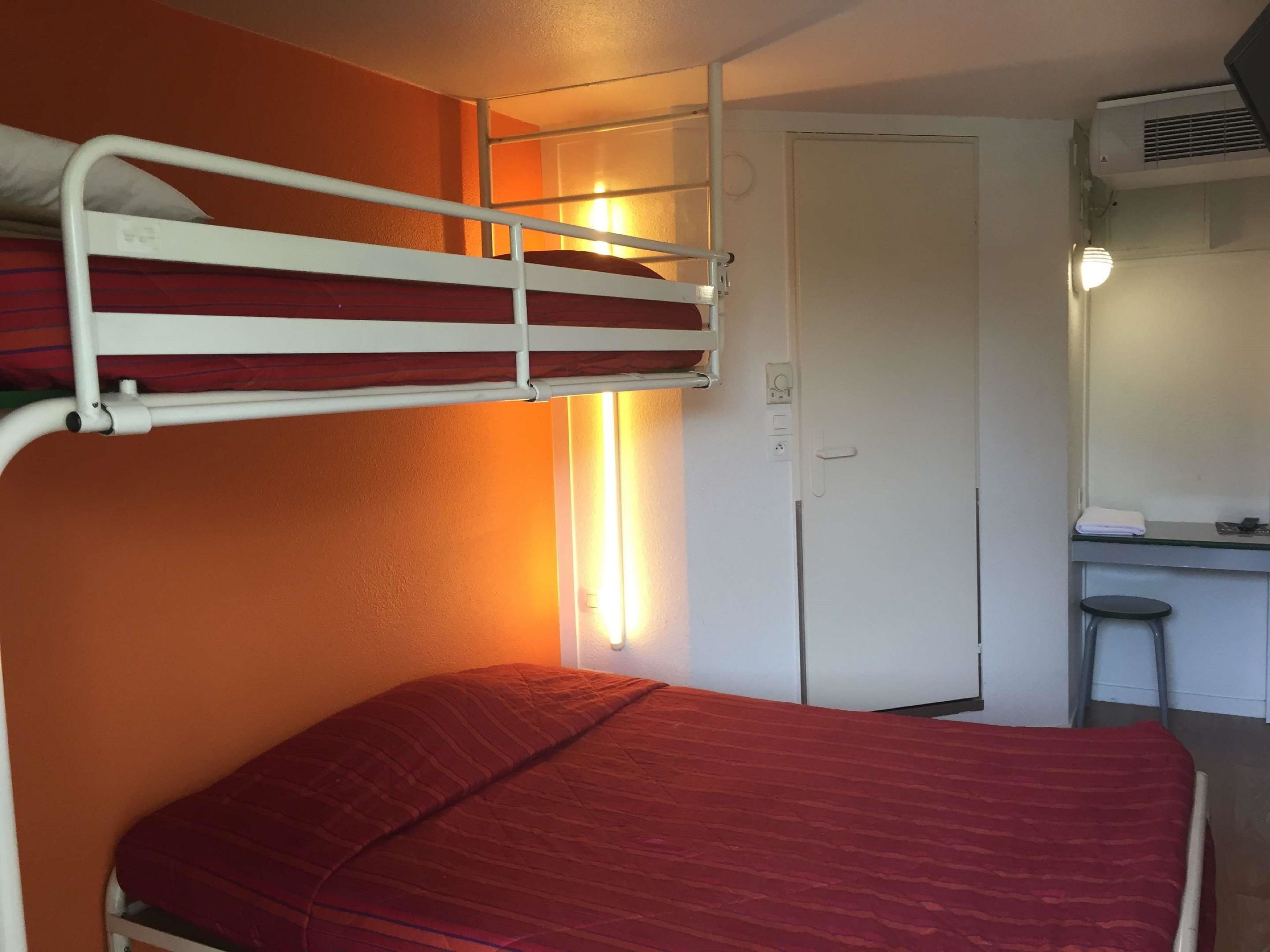 Uma cama beliche com colchão vermelho, uma escrivaninha com cadeira e uma porta branca.