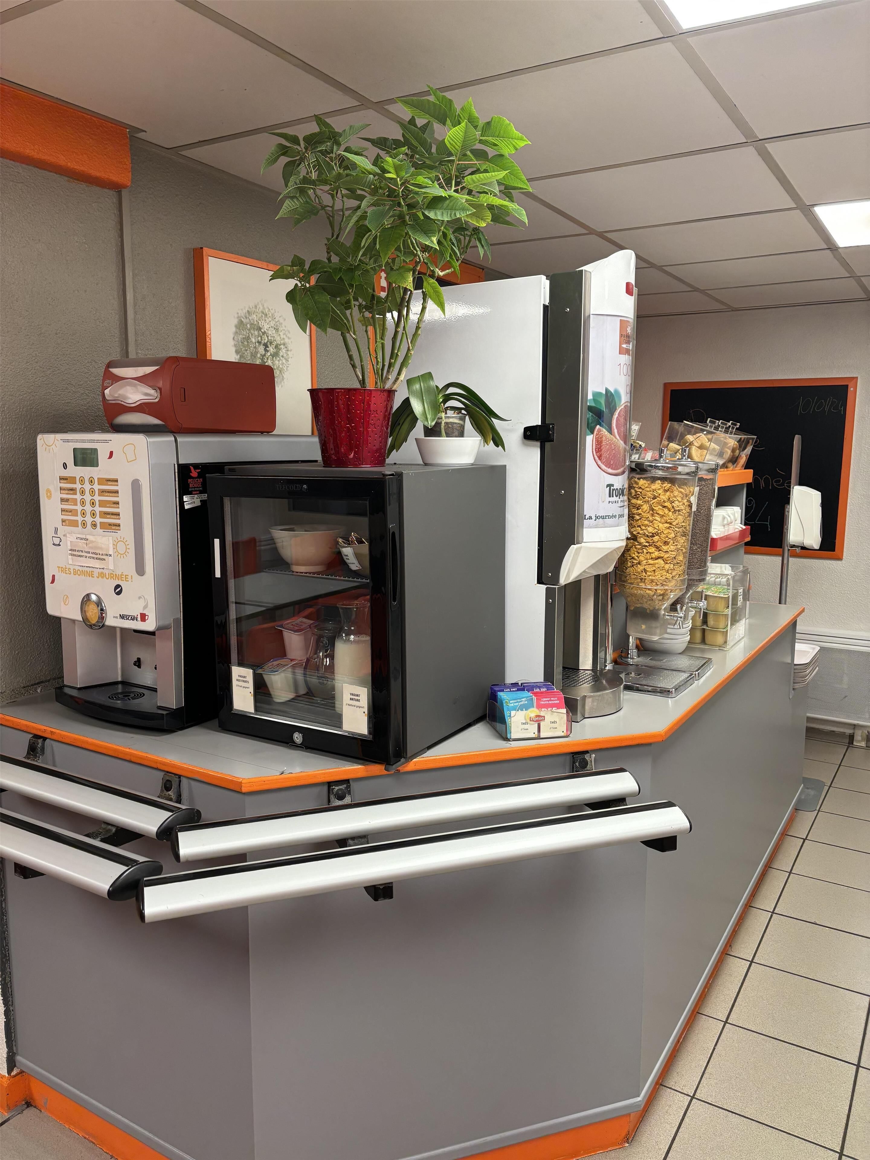 Un espace café en libre-service équipé d’une machine à café Nescafé, d’un réfrigérateur, d’une plante en pot et de divers produits alimentaires.