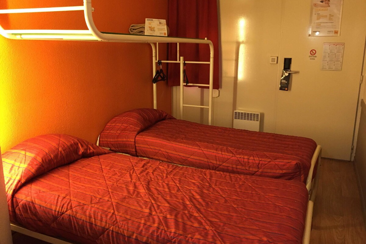 Une petite chambre avec un lit simple, une couverture rouge, un mur blanc, un rideau rouge et une étagère blanche sur laquelle est posé un petit objet.