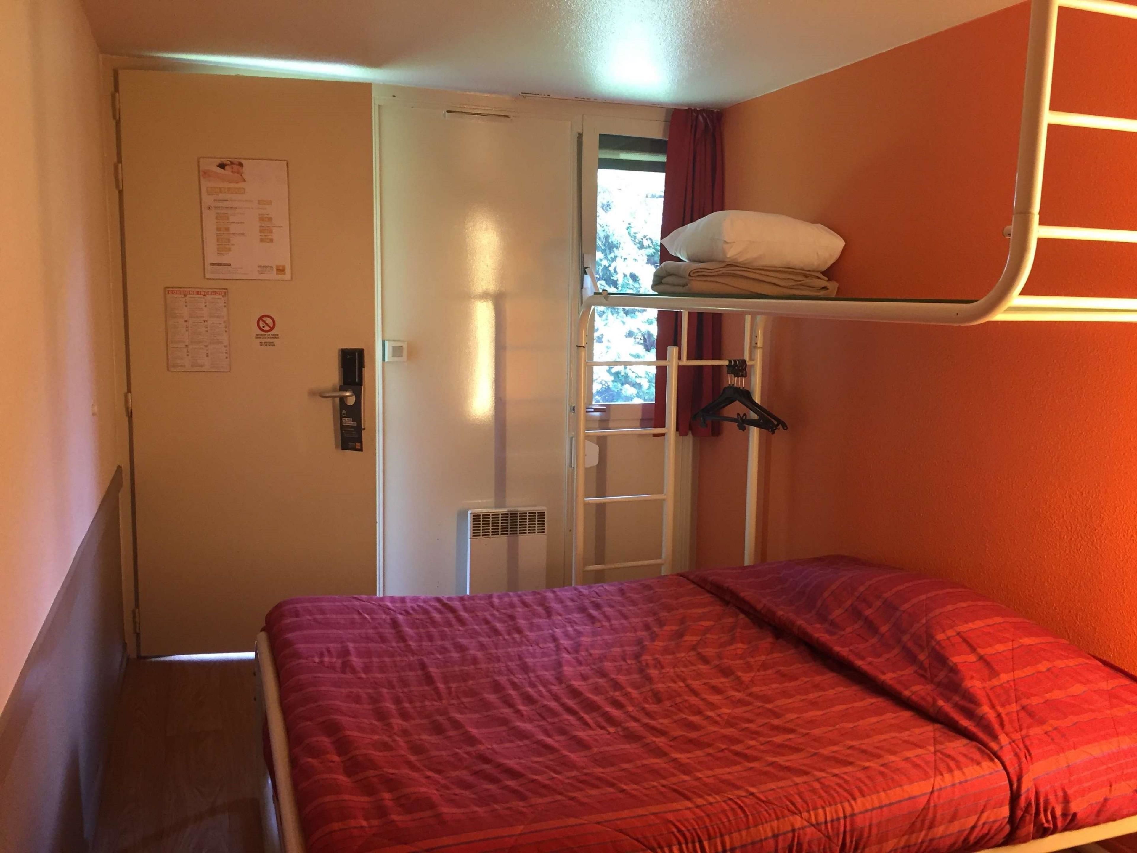 Quarto com beliche, colcha vermelha, porta com aviso, janela com cortinas e um cabideiro.