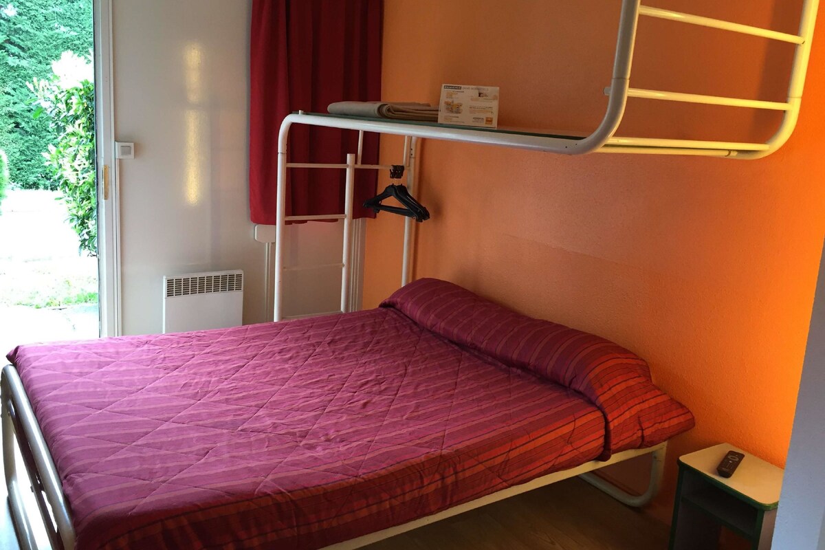 Un lit simple avec une couette rouge, un lit superposé avec un rideau rouge, une petite table avec une télécommande et une fenêtre donnant sur un paysage verdoyant.