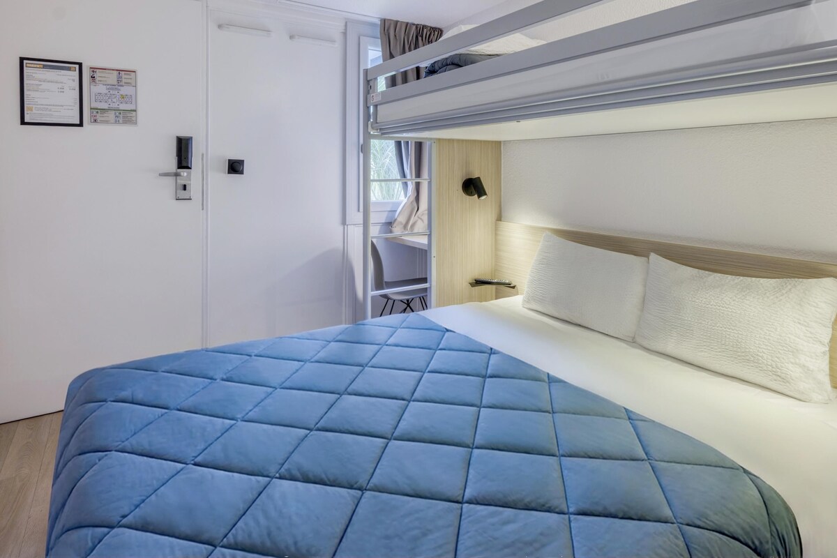 Ein Hotelzimmer mit Etagenbett, einem Bett mit blauer Steppdecke, einem hölzernen Kopfteil und einem kleinen Schreibtisch mit Lampe.