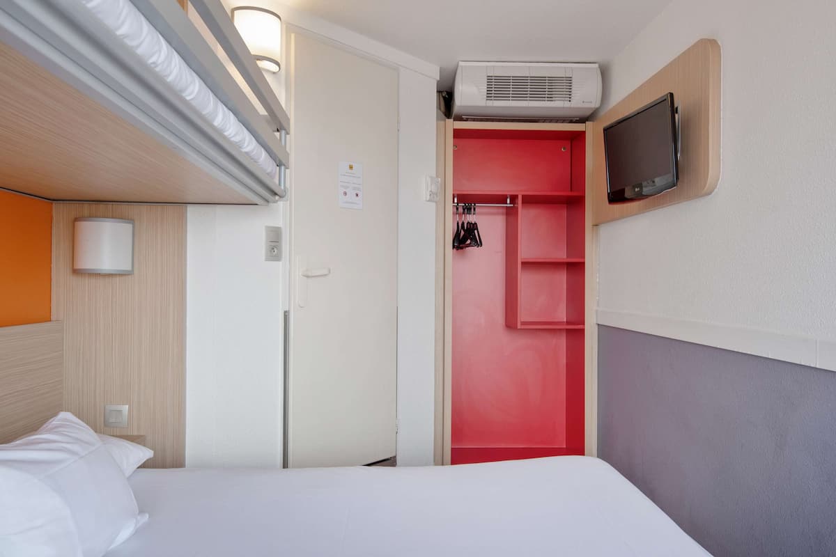 Quarto de hotel com cama, guarda-roupa vermelho, televisão na parede e ar condicionado sobre o guarda-roupa.