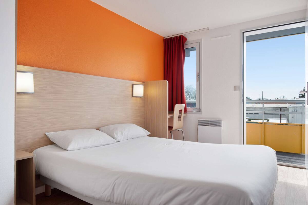 Quarto de hotel com cama, escrivaninha, cadeira e vista para o exterior.
