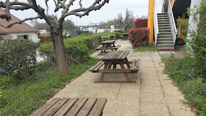 Un sentier dallé, agrémenté de tables de pique-nique en bois, entouré de verdure et bordé d’un bâtiment doté d’un escalier.