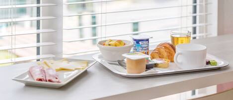 Bandeja de desayuno con jamón, queso, yogur, fruta, croissant y café sobre una mesa junto a una ventana con persianas.