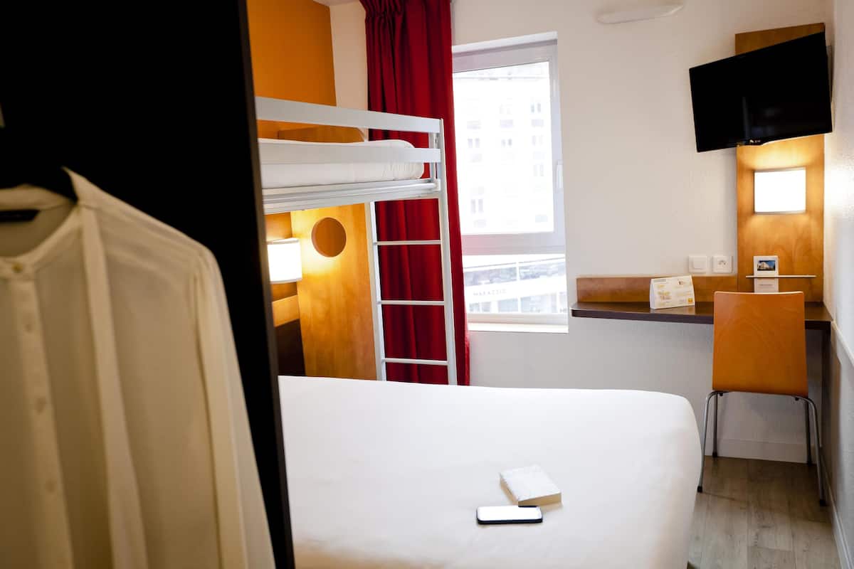 Une chambre d’hôtel avec un lit superposé, un bureau avec une chaise, un téléviseur fixé au mur et une fenêtre donnant sur des immeubles.