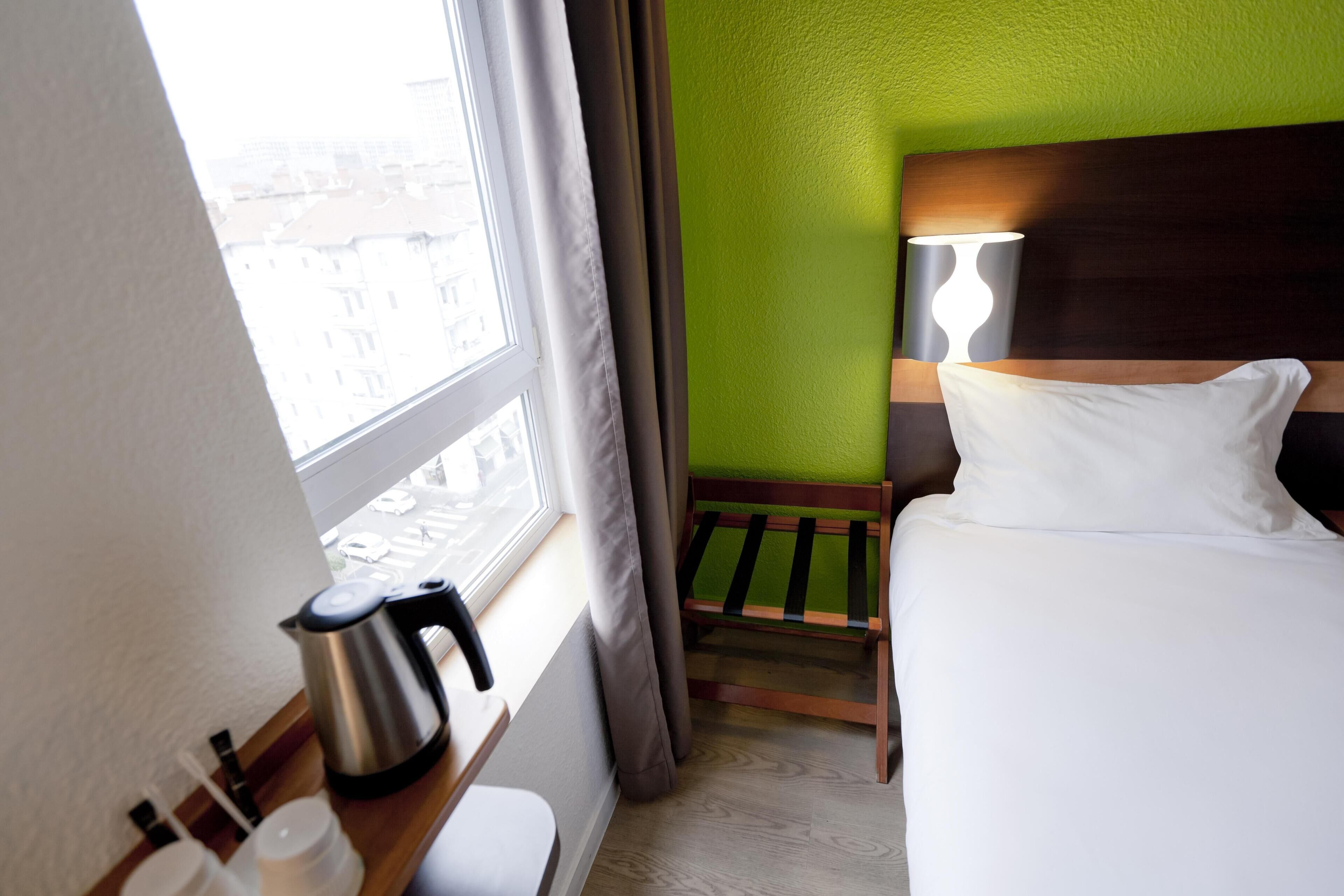 A hotel room with a bed, a nightstand, a kettle, and a window with curtains.