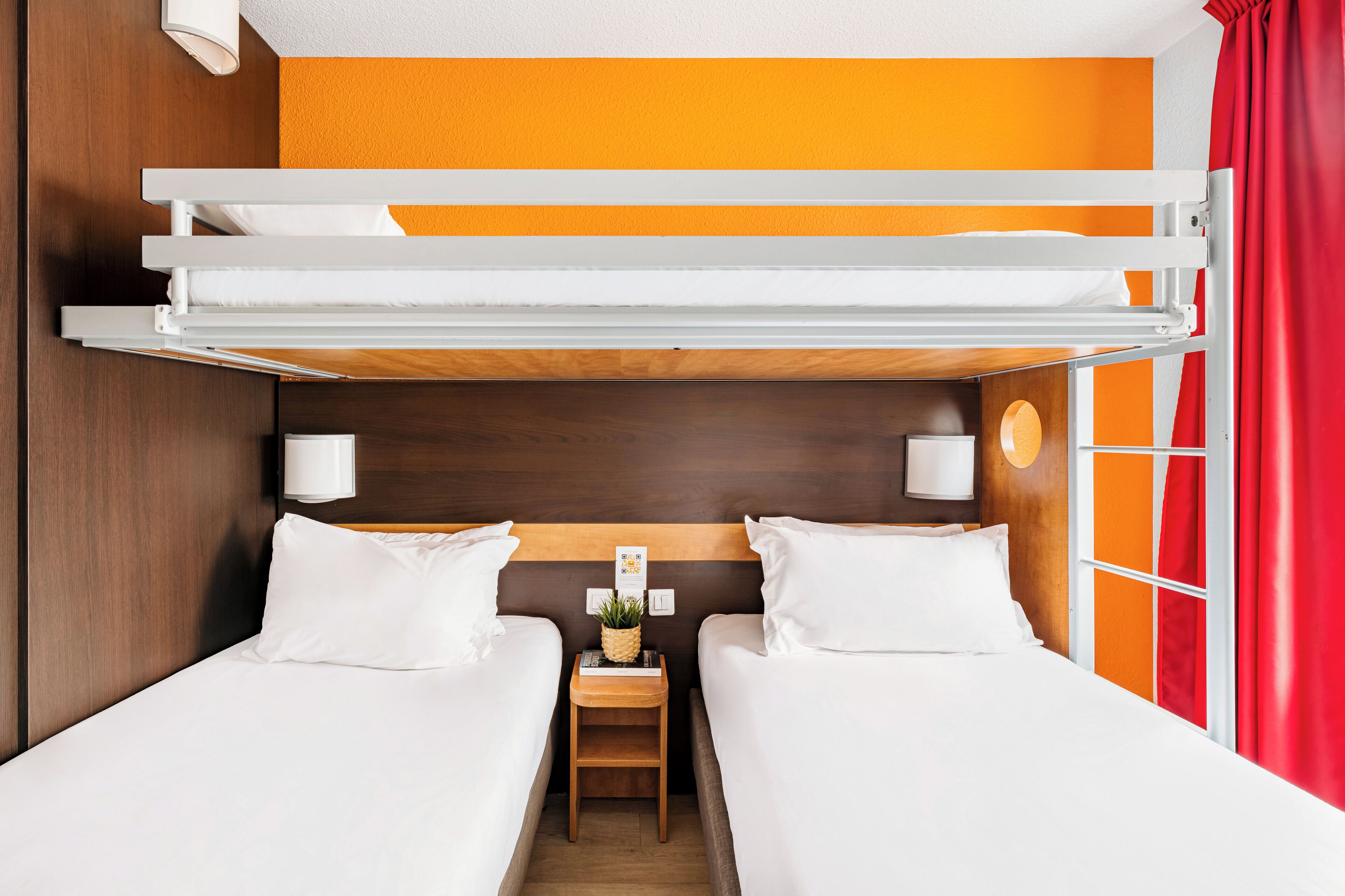 A bunk bed room with two beds, a nightstand with a lamp, and a window with orange curtains.