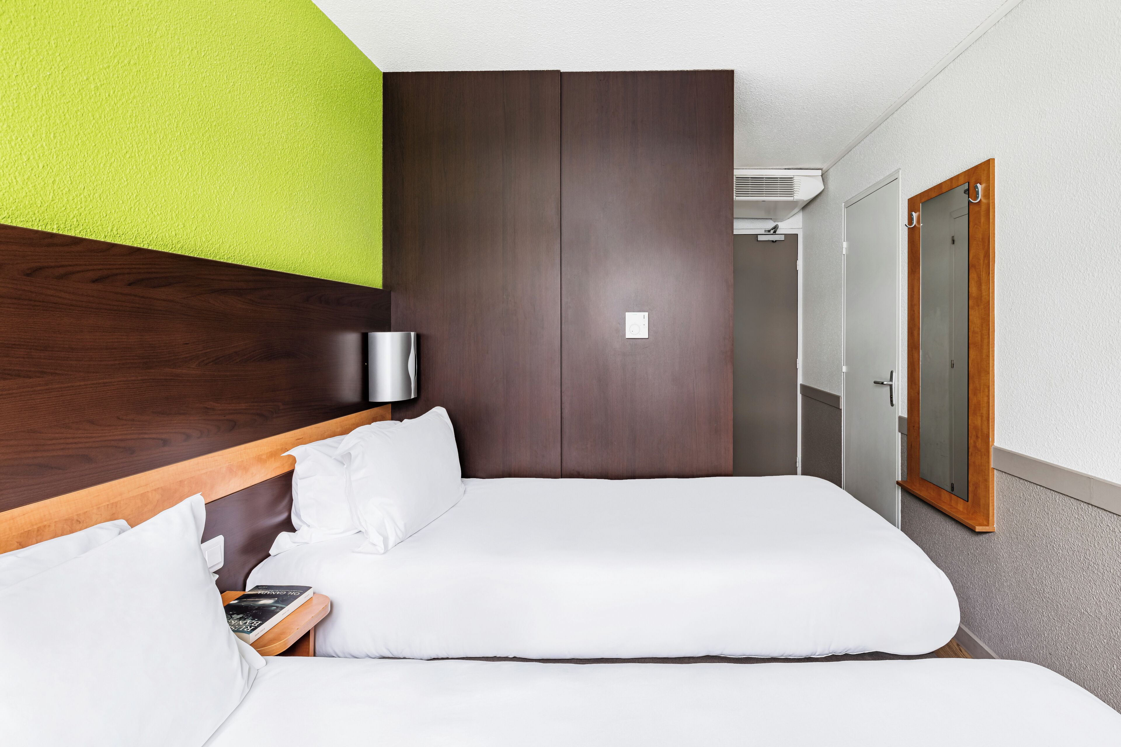 A hotel room with a bed, a bedside table with a lamp, a mirror, and a door to a bathroom.