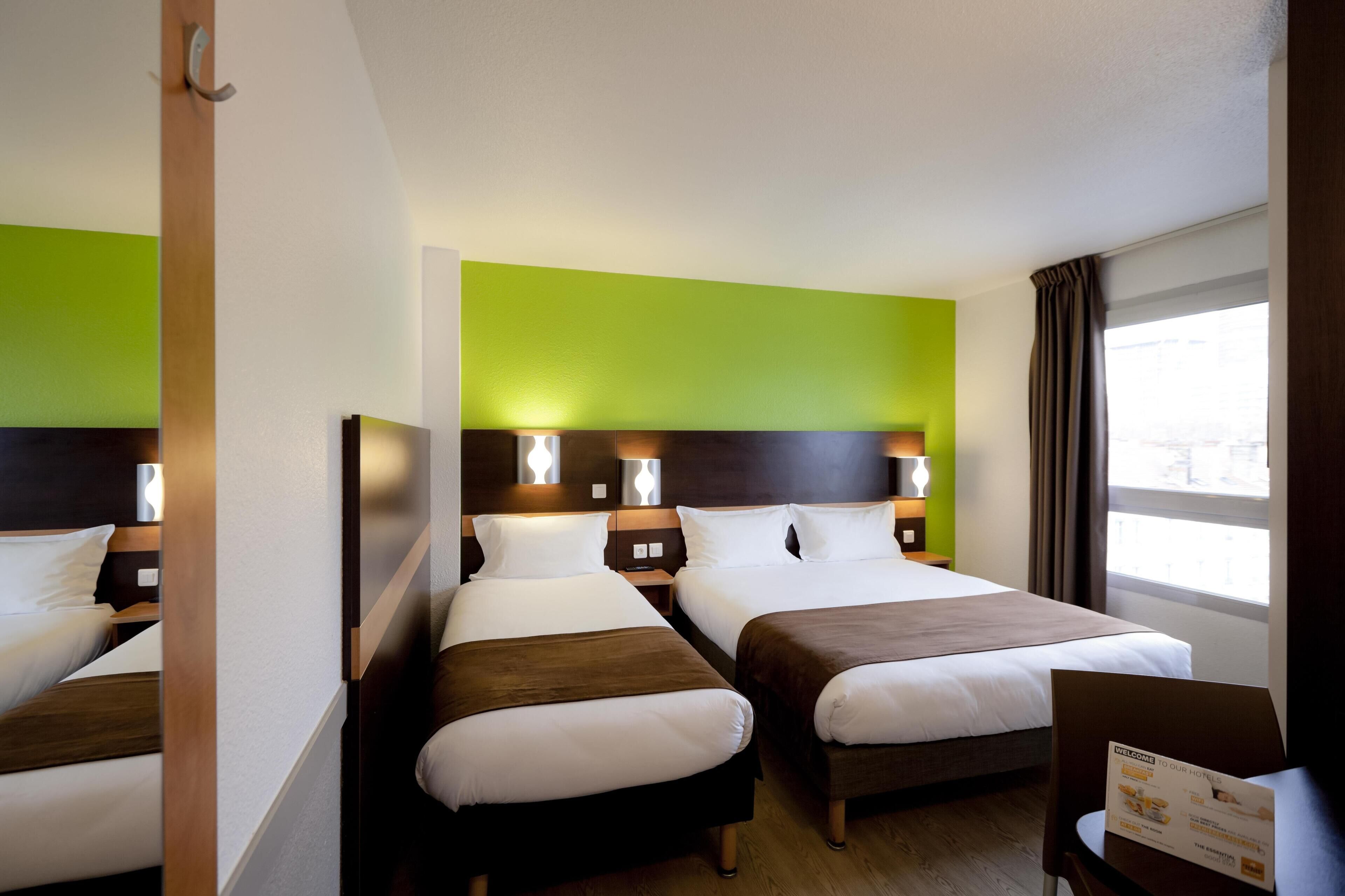 A hotel room with two beds, a green accent wall, bedside lamps, a window with curtains, and a welcome brochure on a table.