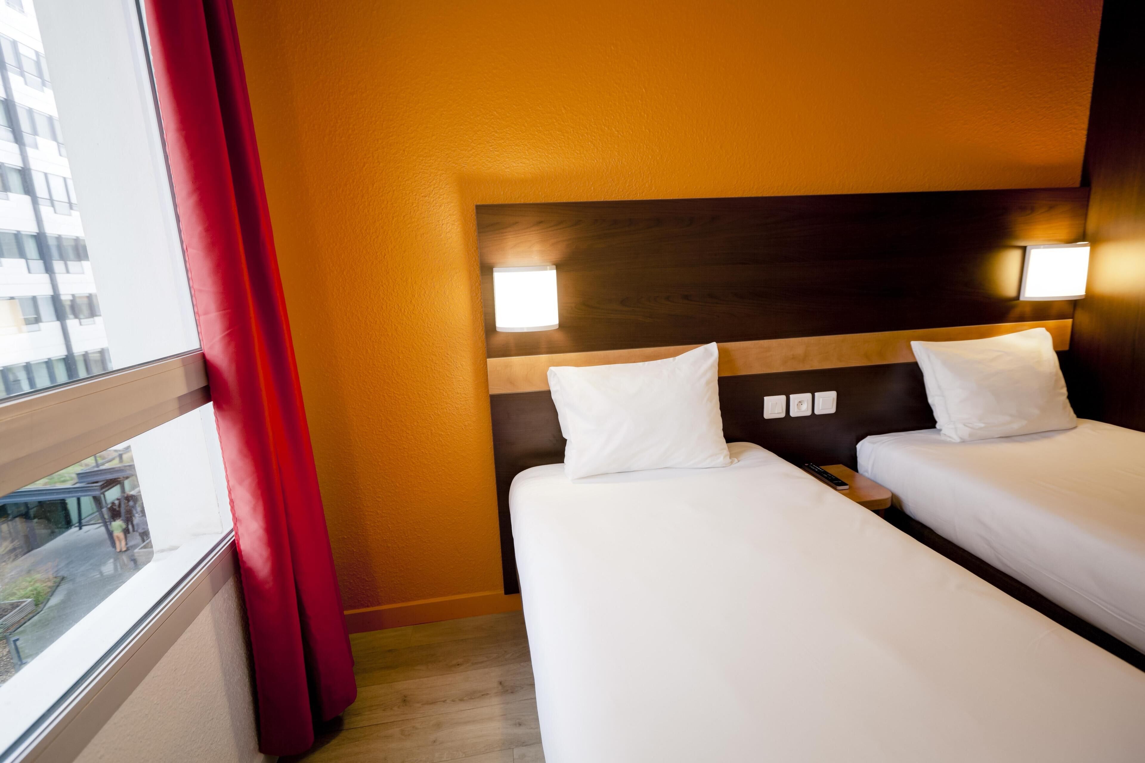 Habitación de hotel con dos camas, cabecero de madera, ventana con cortinas rojas y vista a un edificio.