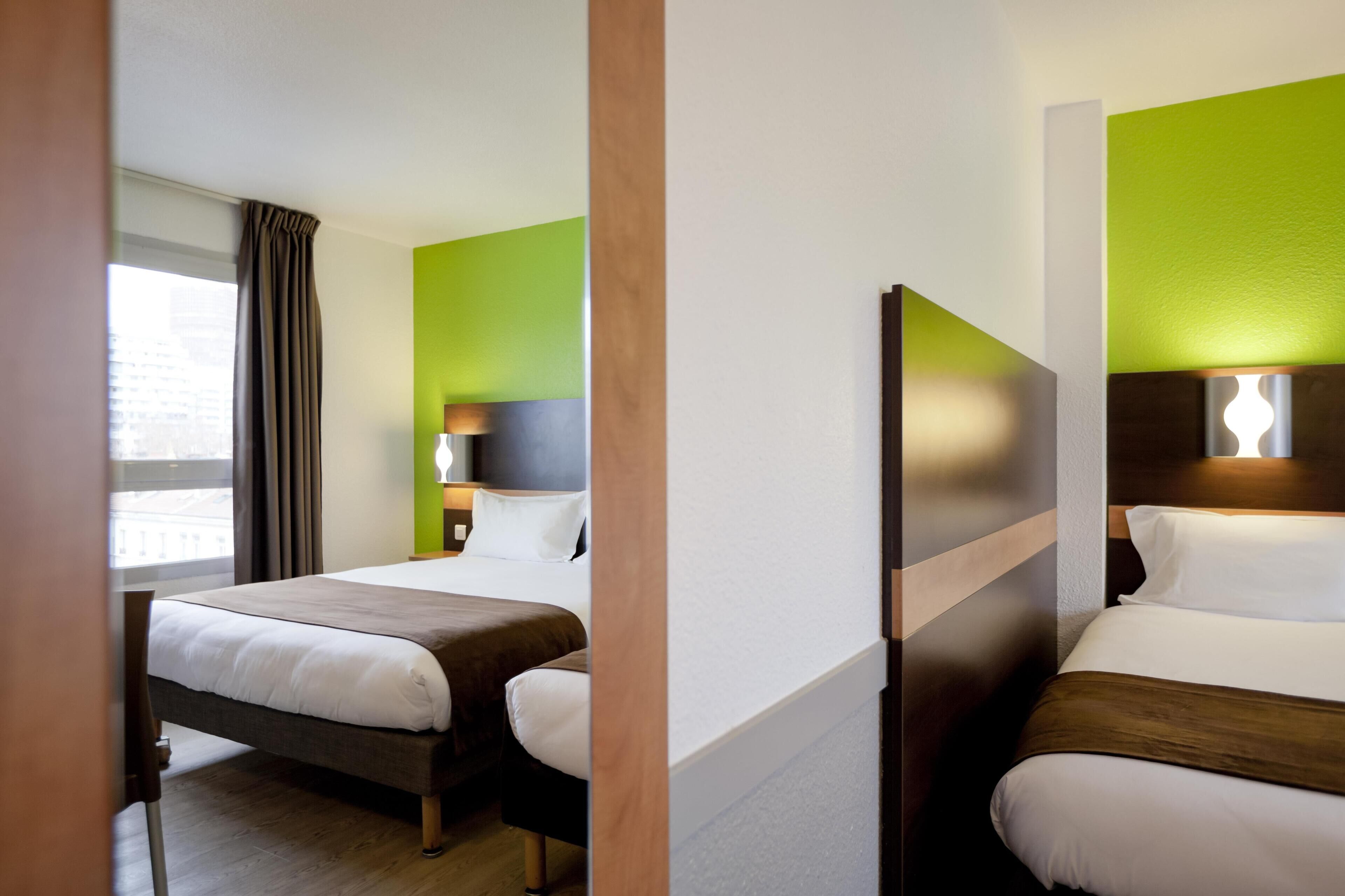 Habitación de hotel con dos camas, una ventana con cortinas, una mesita de noche con lámpara y una pared de acento verde.