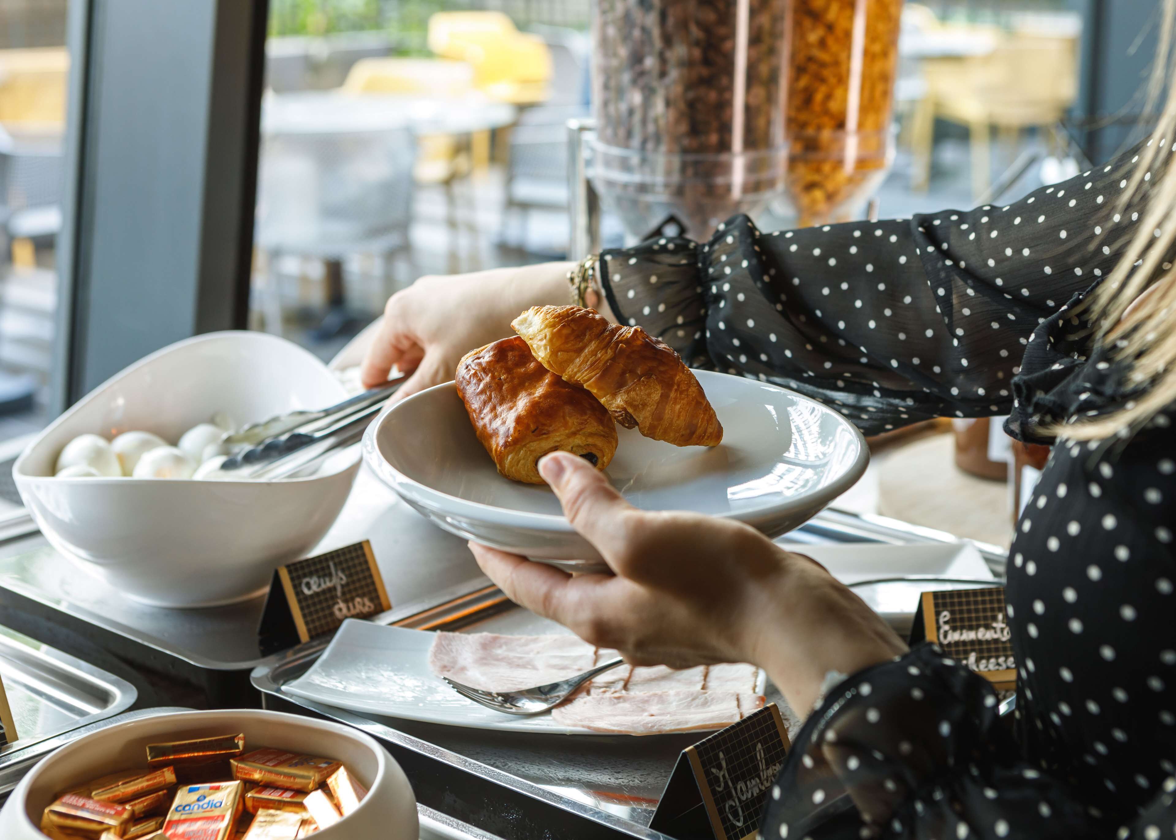 A person holding a plate with croissants and a bowl of eggs, with a menu and condiments on the table.