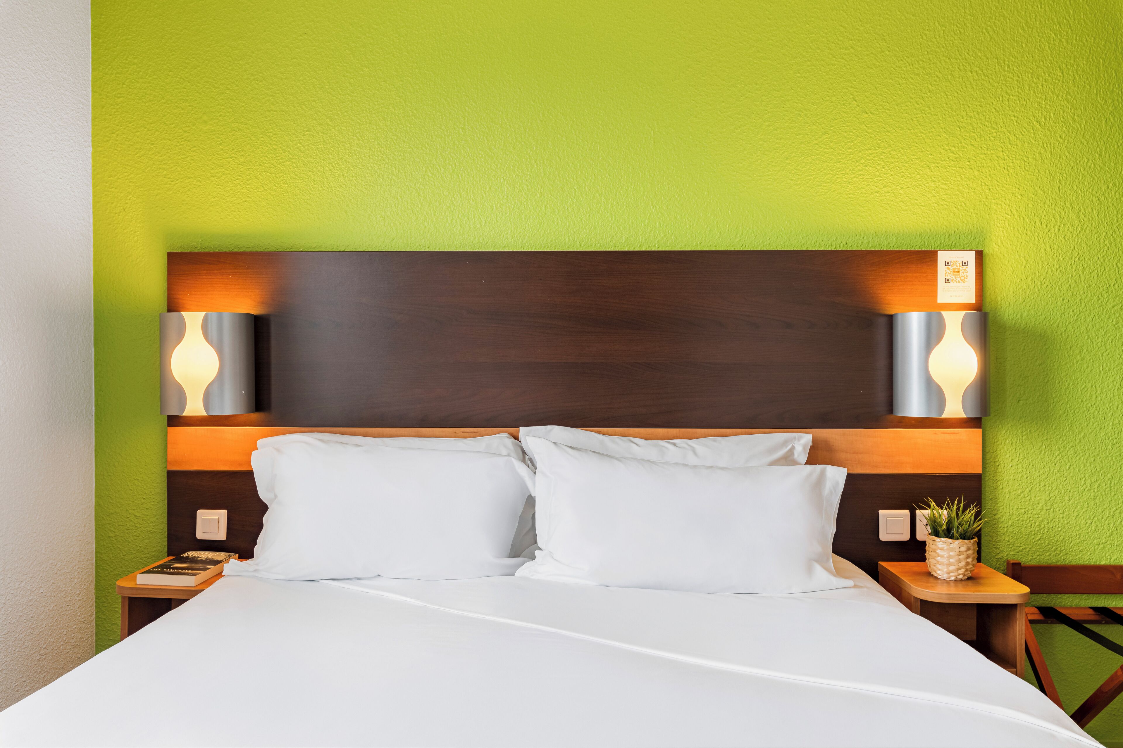 A neatly made bed with white linens, two bedside lamps, and a small bedside table with a plant.