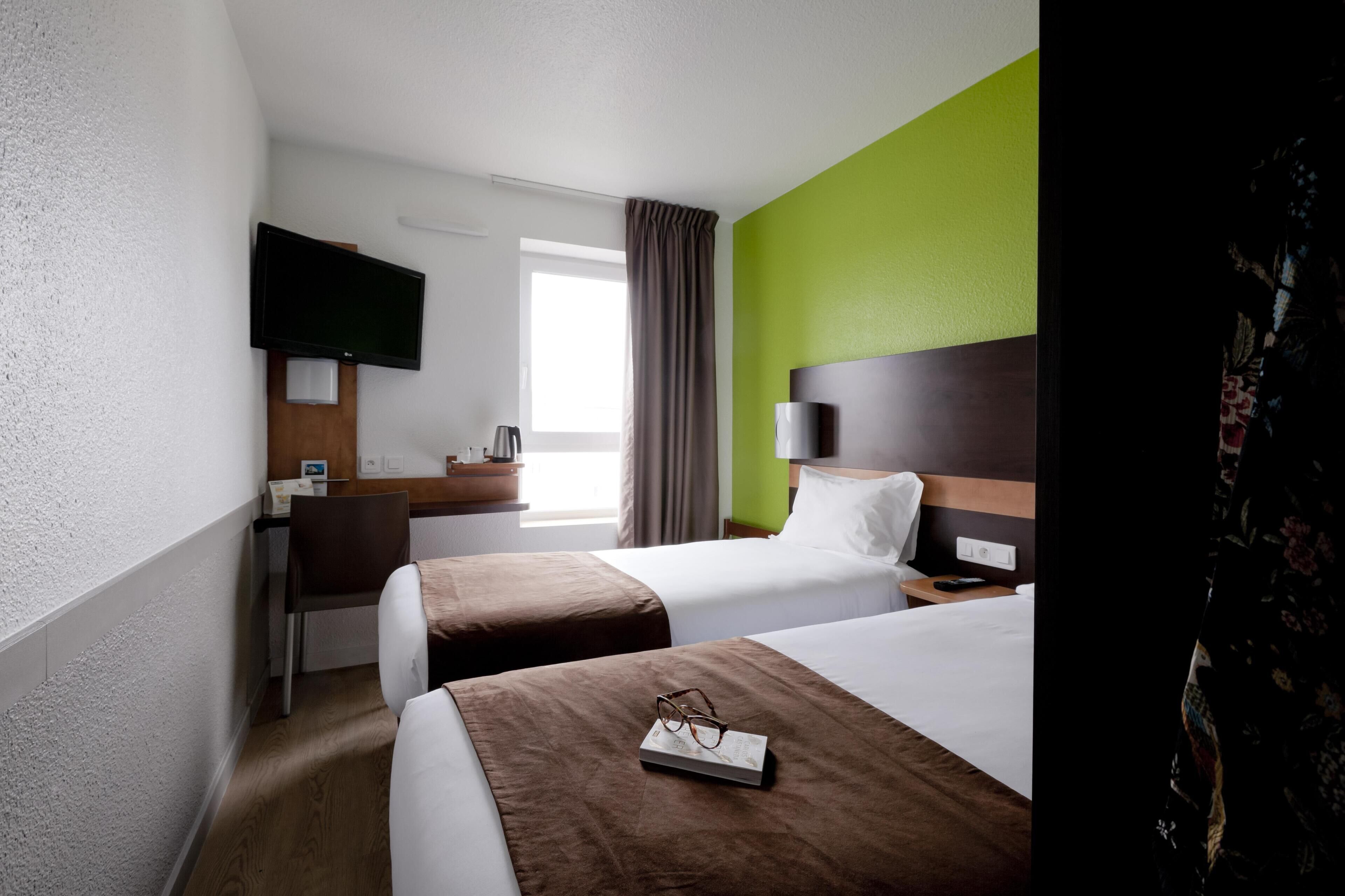 A hotel room with two beds, a desk, a television, and a window with curtains.