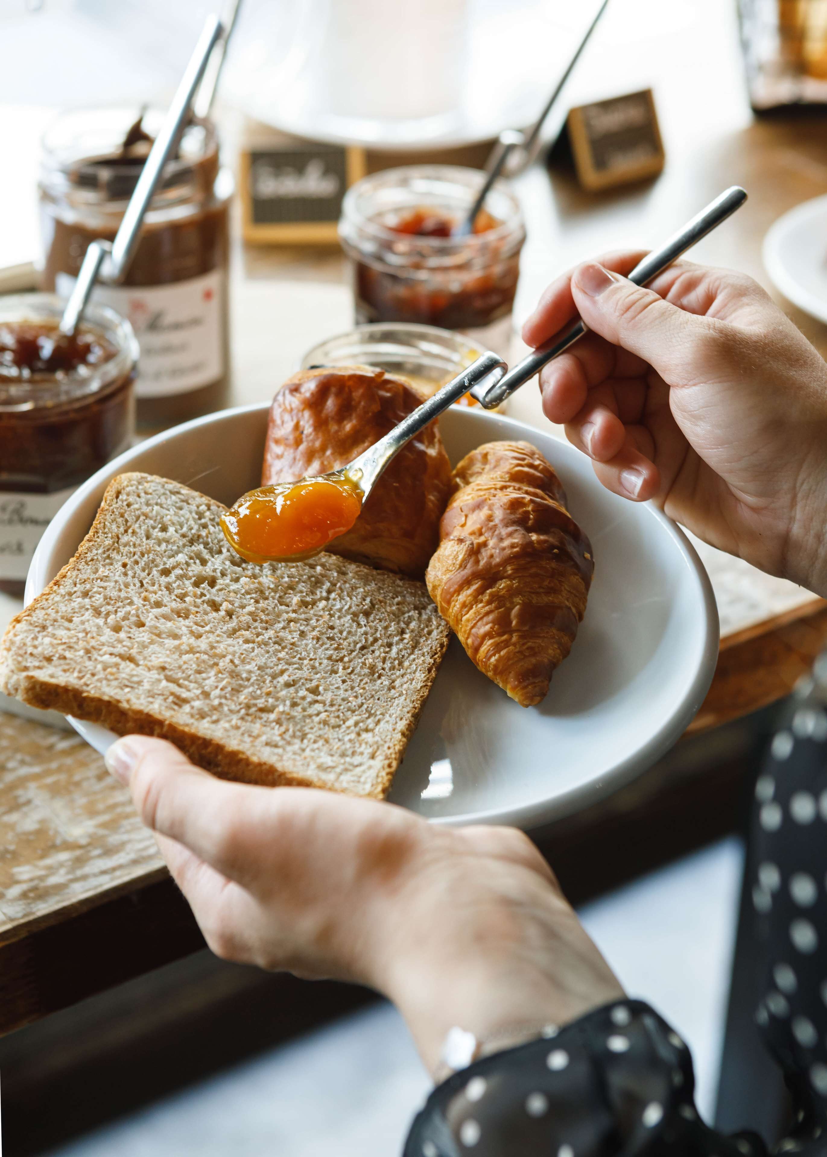 A person spreading jam on a croissant and a slice of bread.