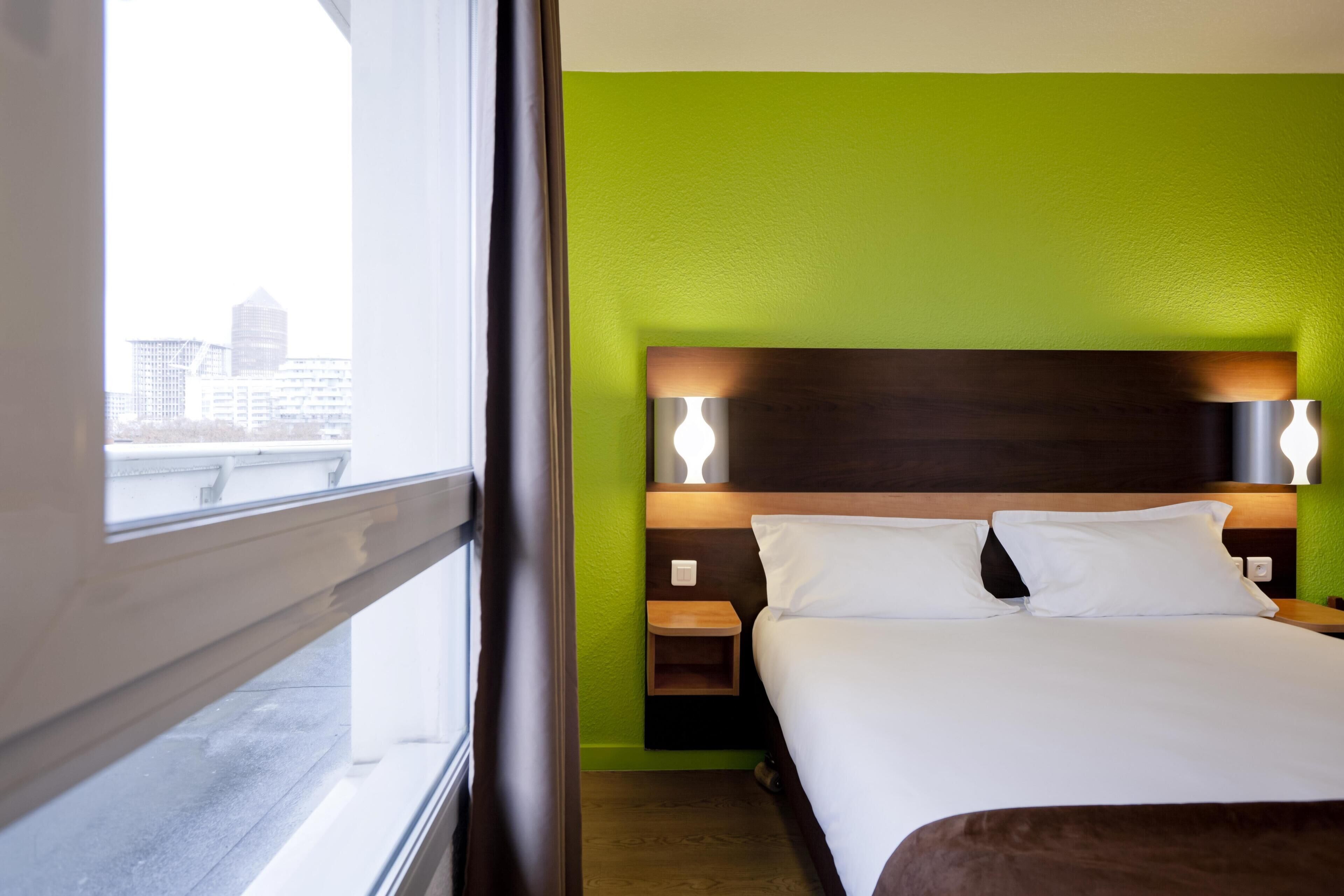 A hotel room with a bed, a nightstand, and a window with a view of buildings.