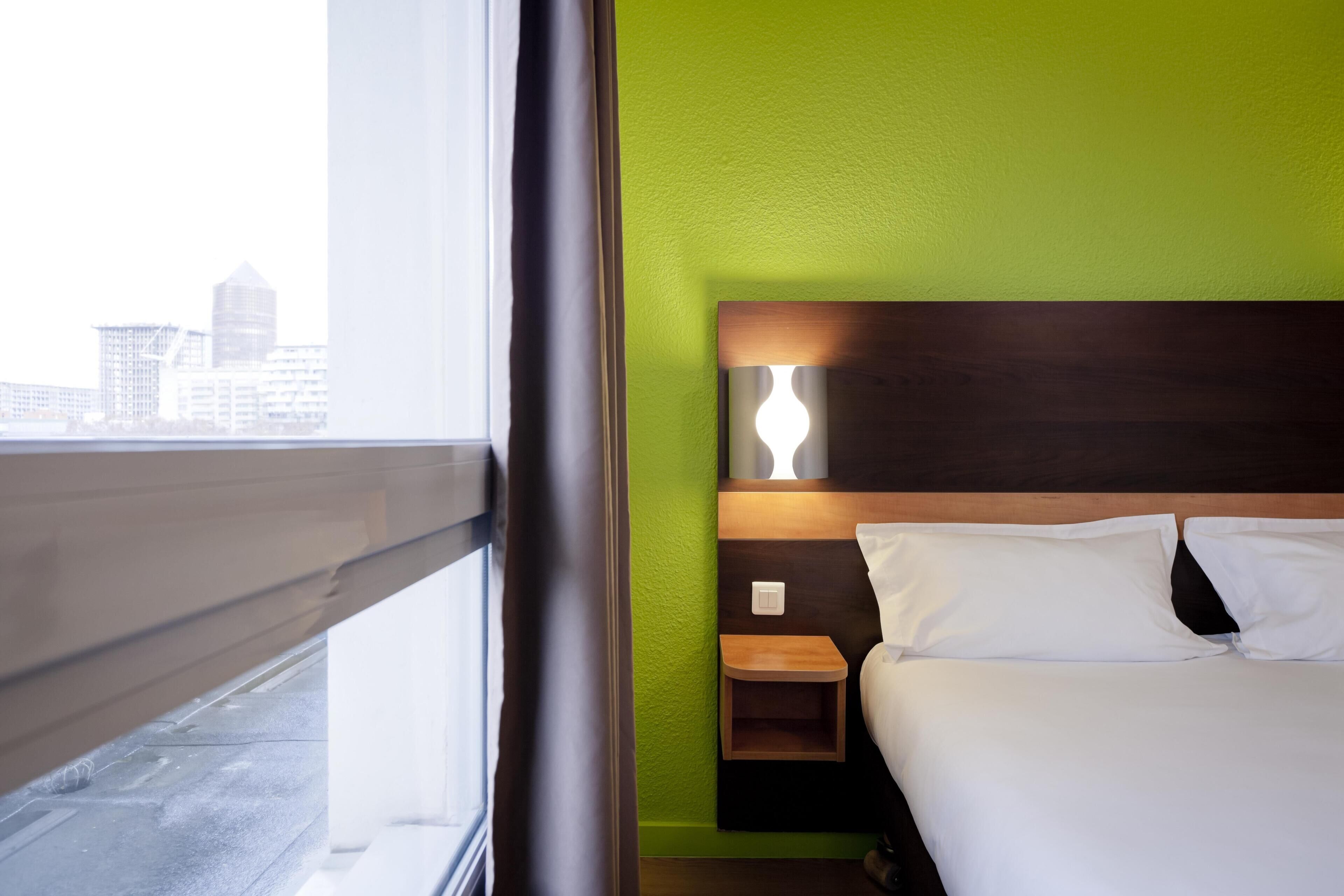 Habitación de hotel con cama, mesita de noche, lámpara de pared y ventana con vista a edificios.