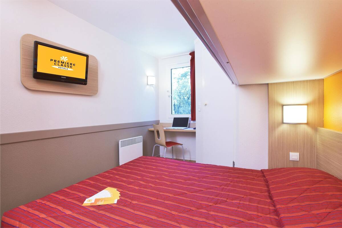 Ein Hotelzimmer mit einem Bett, einem Schreibtisch mit einem Laptop, einem Sessel und einem an der Wand befestigten Fernseher, der "Première Classe Hotels" anzeigt.