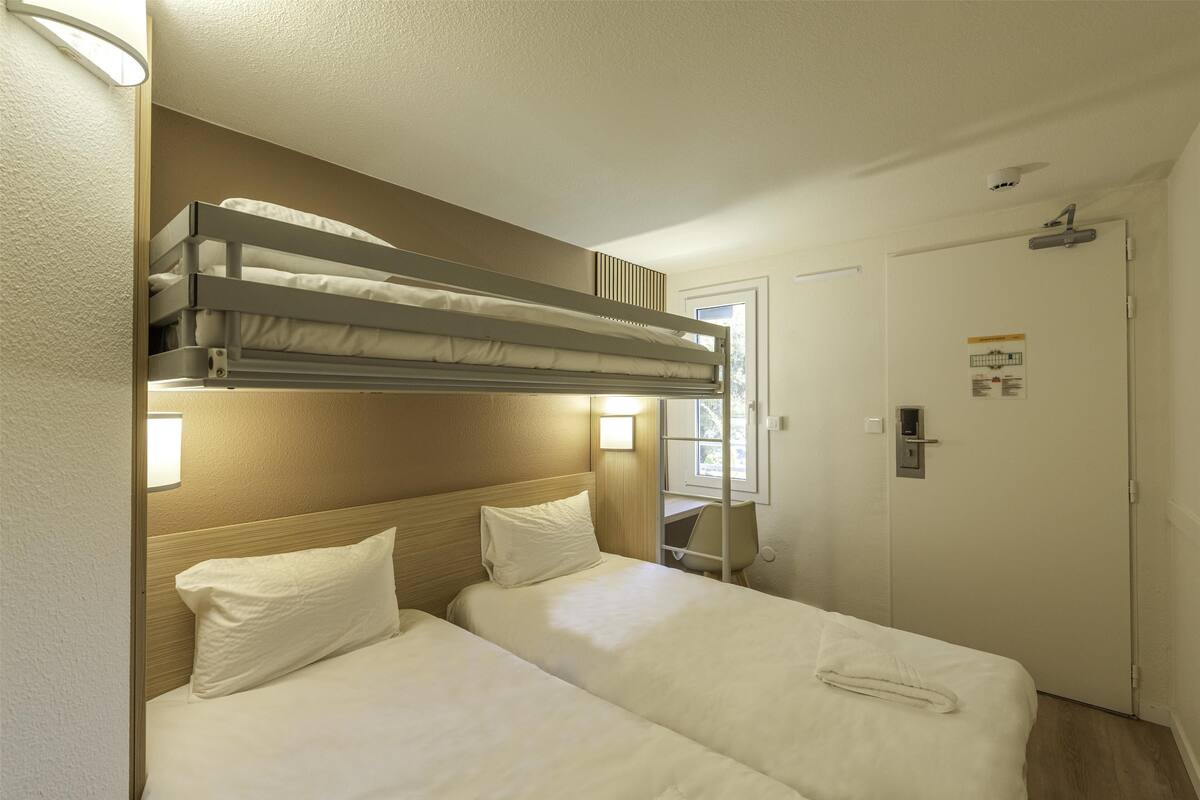 A hotel room with a bunk bed, two single beds, a desk with a chair, a door, and a window.