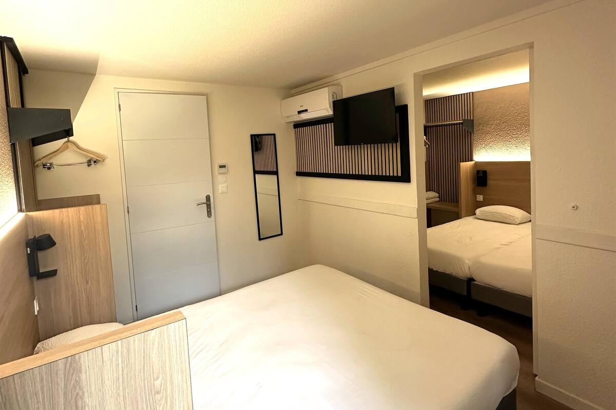 Una habitación de hotel moderna con una cama grande, un televisor de pantalla plana montado en la pared, un equipo de aire acondicionado integrado y una pequeña zona de estar con un banco de madera.