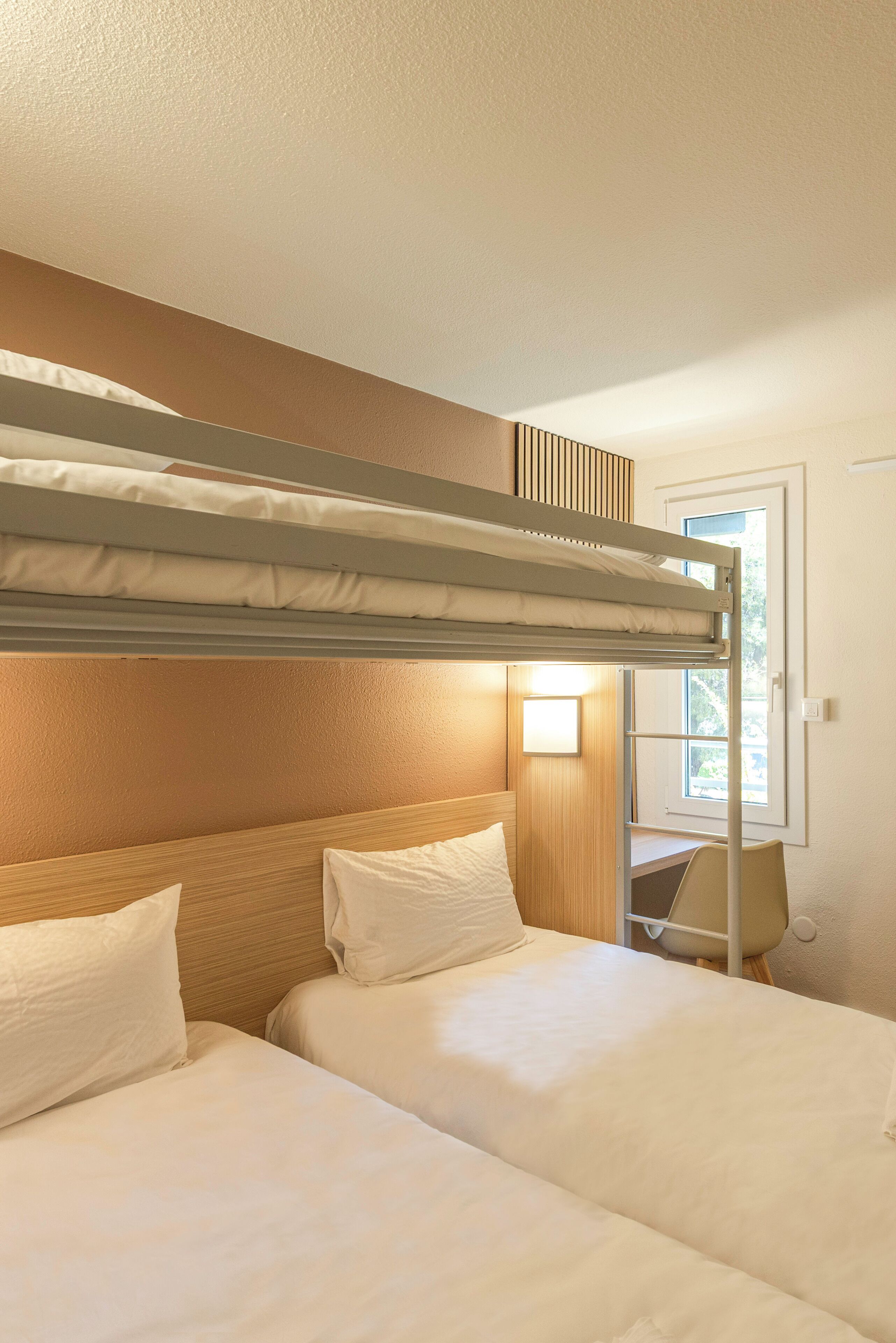 Ein Stockbett mit zwei Betten, einem Schreibtisch, einem Stuhl und einem Fenster mit Tageslicht.