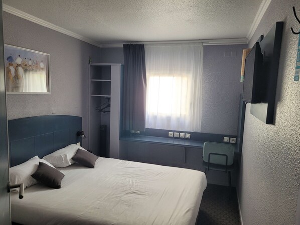 Habitación de hotel con una cama, un escritorio, una silla, una ventana con cortinas y un cuadro en la pared.