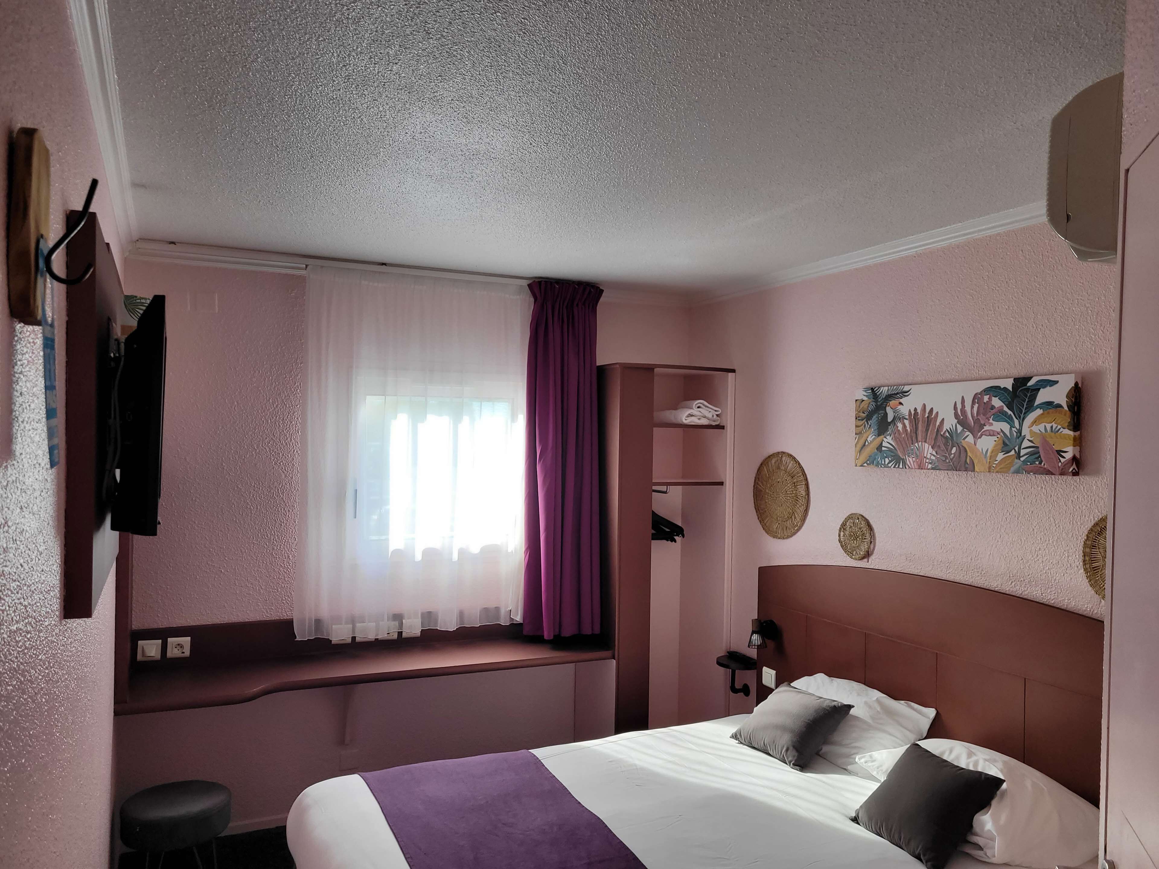 A hotel room with a bed, a window with sheer curtains, a wall-mounted shelf, and a tropical-themed painting.