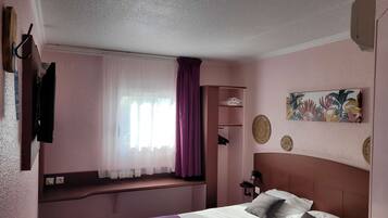 Quarto de hotel com cama, janela com cortinas leves, prateleira na parede e pintura com tema tropical.