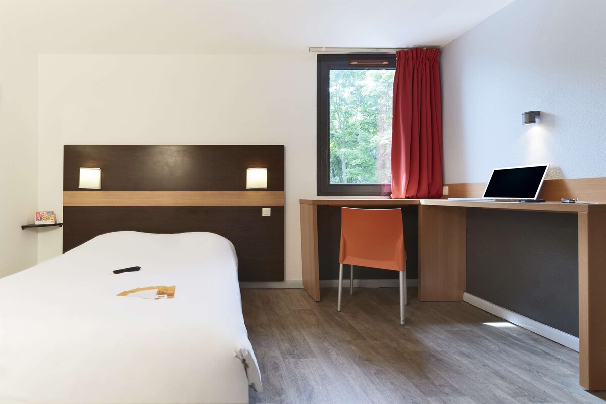 A hotel room with a bed, a desk with a laptop, a chair, and a window with curtains.