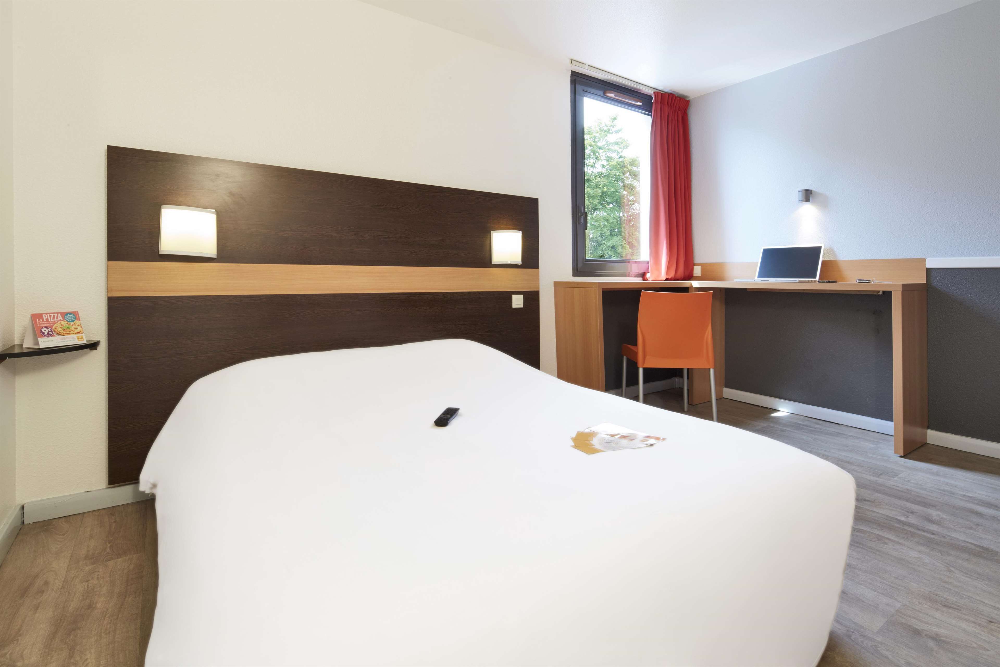 A hotel room with a bed, a desk with a laptop, a chair, and a window with a red curtain.