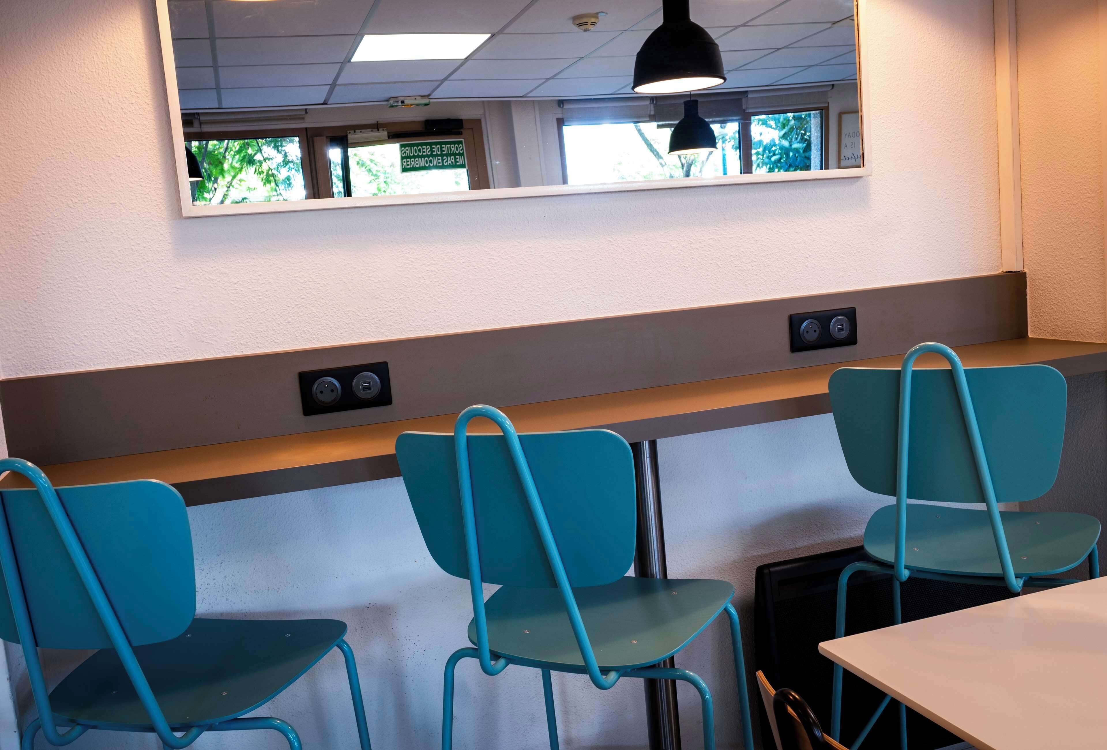 Una zona de estar con sillas color turquesa y una mesa blanca, una ventana con vistas a árboles, y un accesorio de iluminación en el techo.