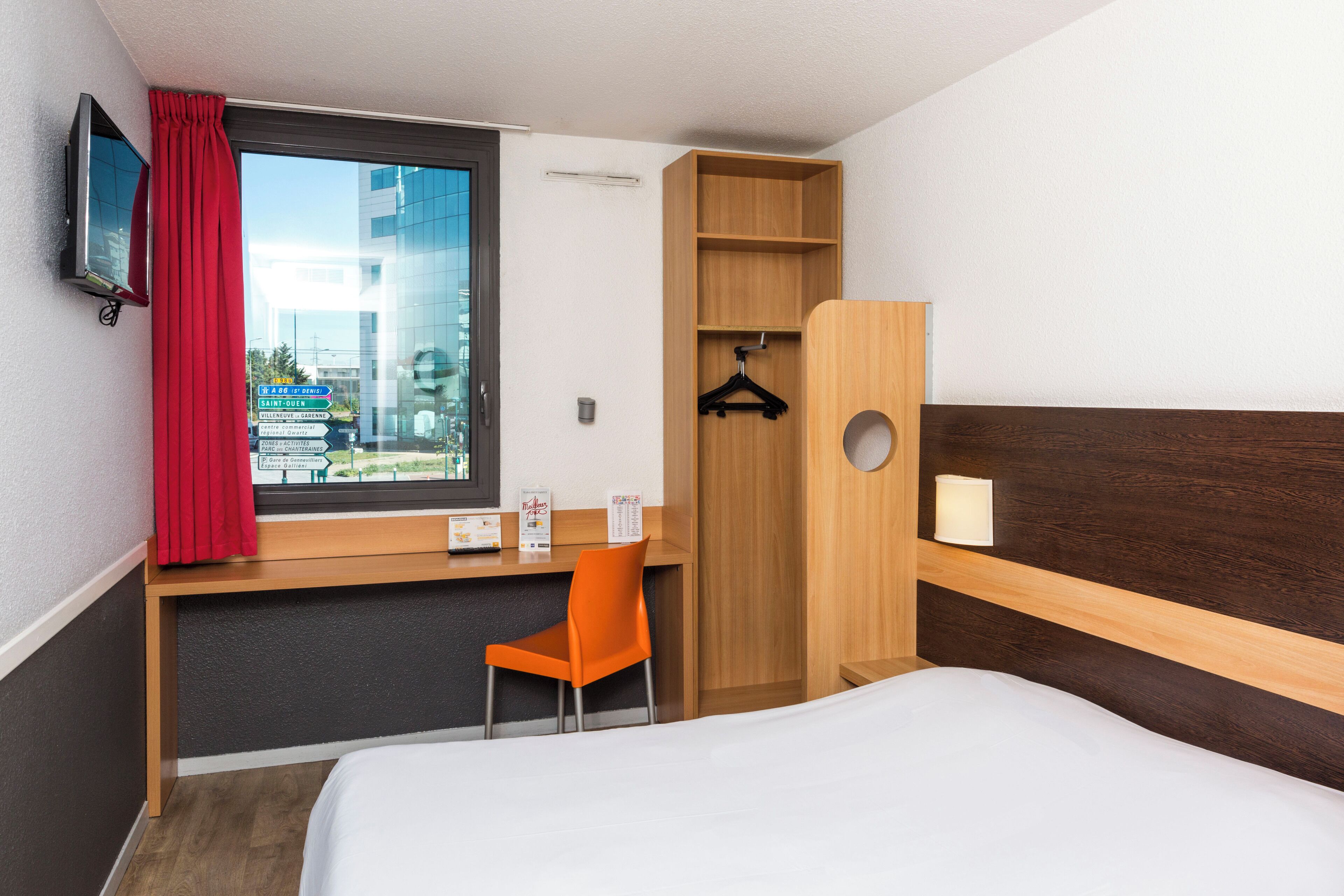 Une chambre d’hôtel comprenant un lit, un bureau avec une chaise, une armoire, une télévision et une fenêtre donnant sur des immeubles.