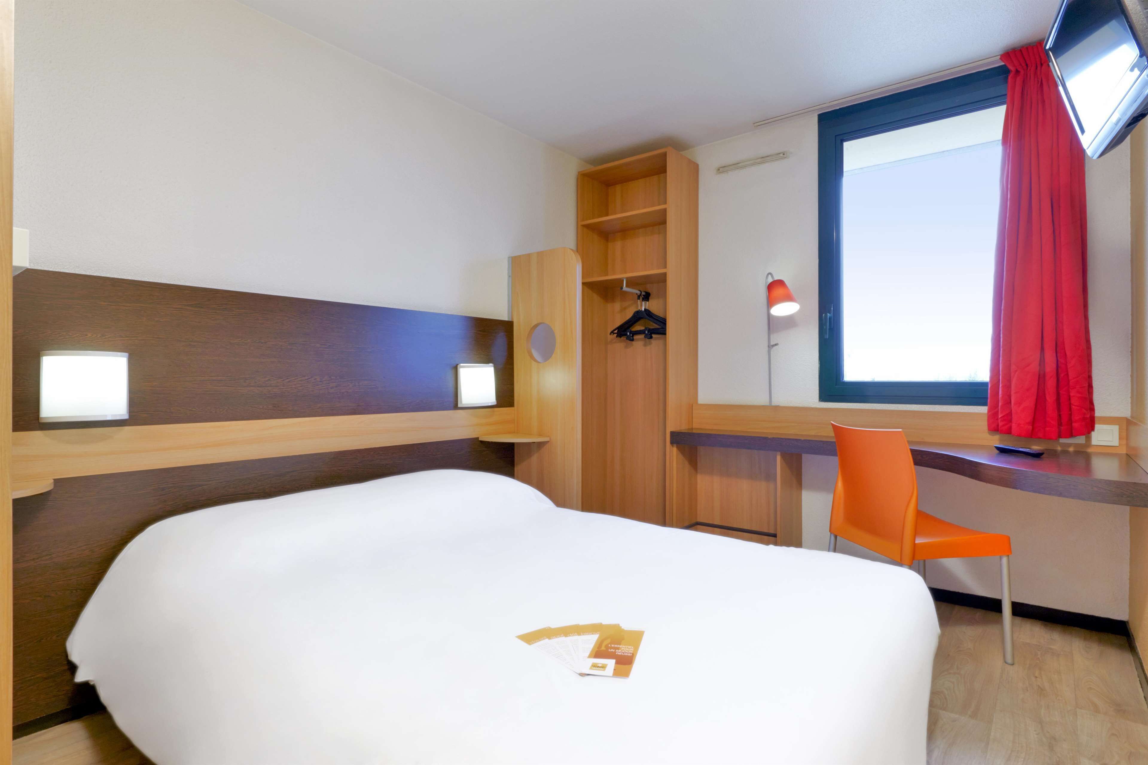 A hotel room with a large bed, a desk with a chair, a wardrobe, and a window with a red curtain.