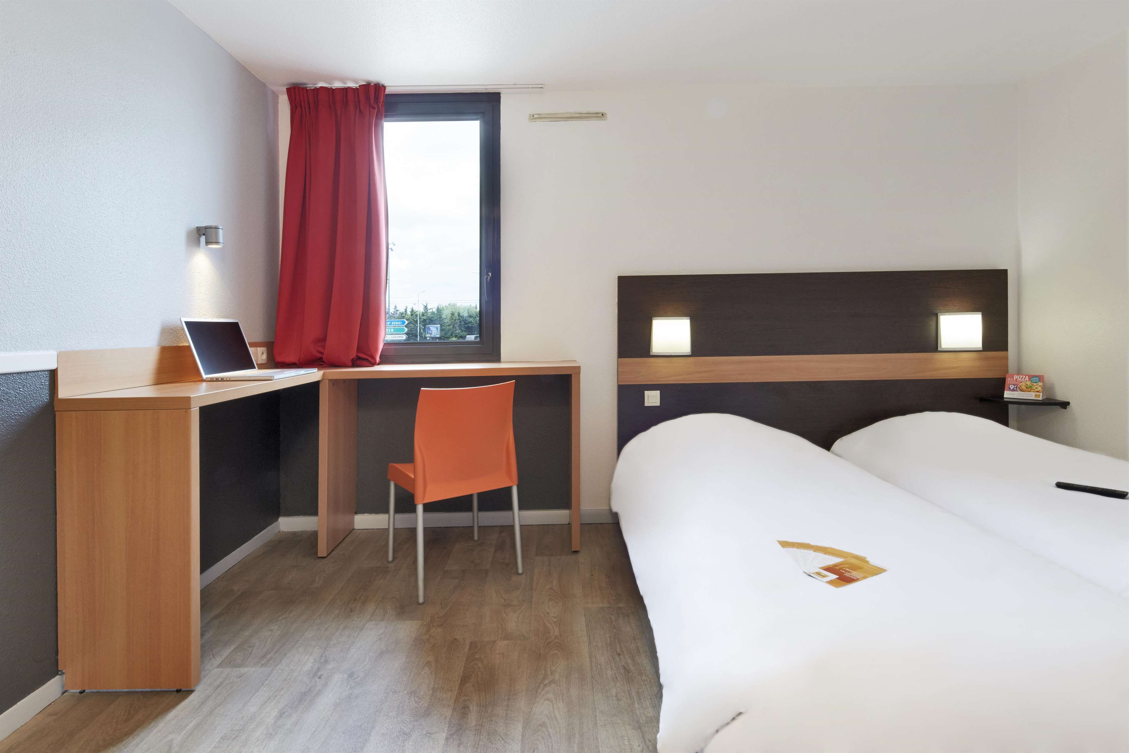 A hotel room with a wooden desk, a laptop, an orange chair, a bed with white bedding, and a window with red curtains.