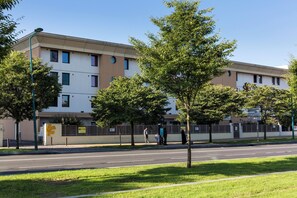 A multi-story building with a flat roof, surrounded by trees and a fence. - Première Classe Paris Ouest - Gennevilliers Barbanniers (Gennevilliers)