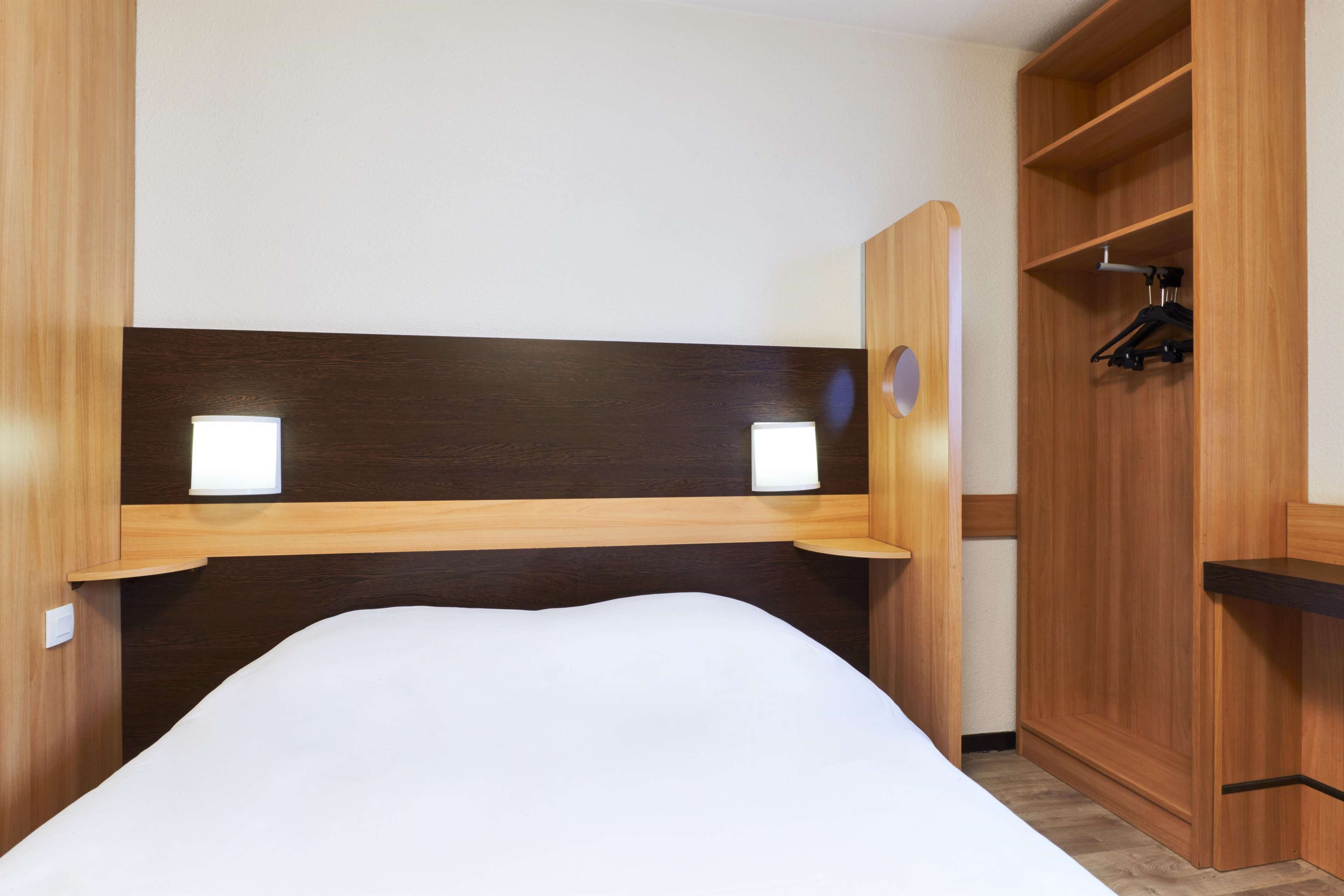 Een hotelkamer met een houten hoofdeinde, twee nachtkastjes met lampen, een kledingkast, en een bed met wit beddengoed.