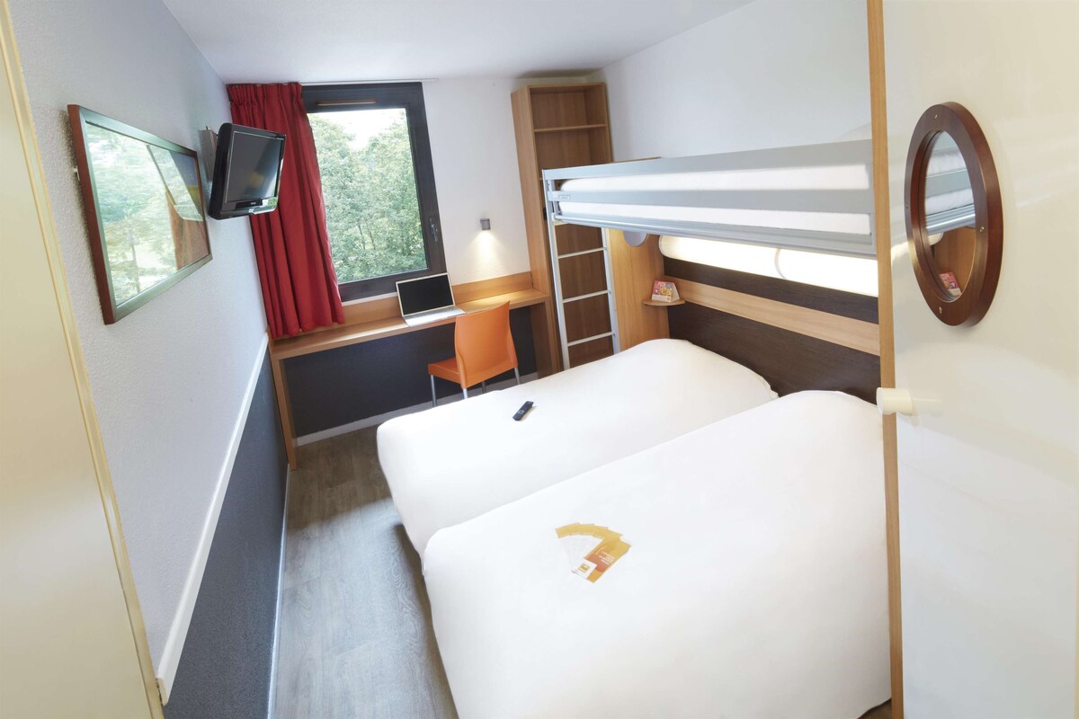 A compact hotel room with a bunk bed, a desk with a laptop, a TV mounted on the wall, and a window with red curtains.