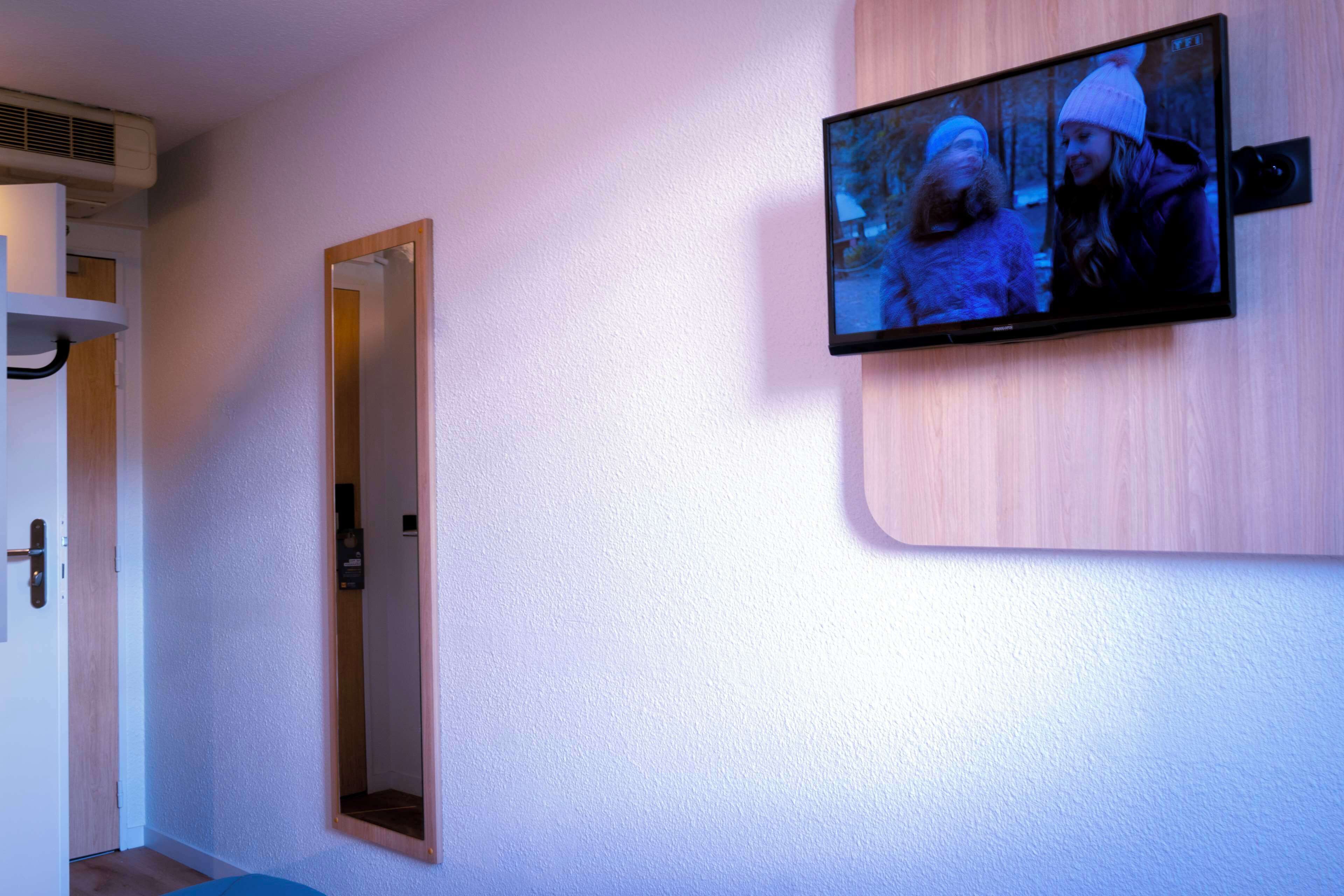 Una habitación con una televisión montada que muestra una escena, una puerta y un espejo en la pared.