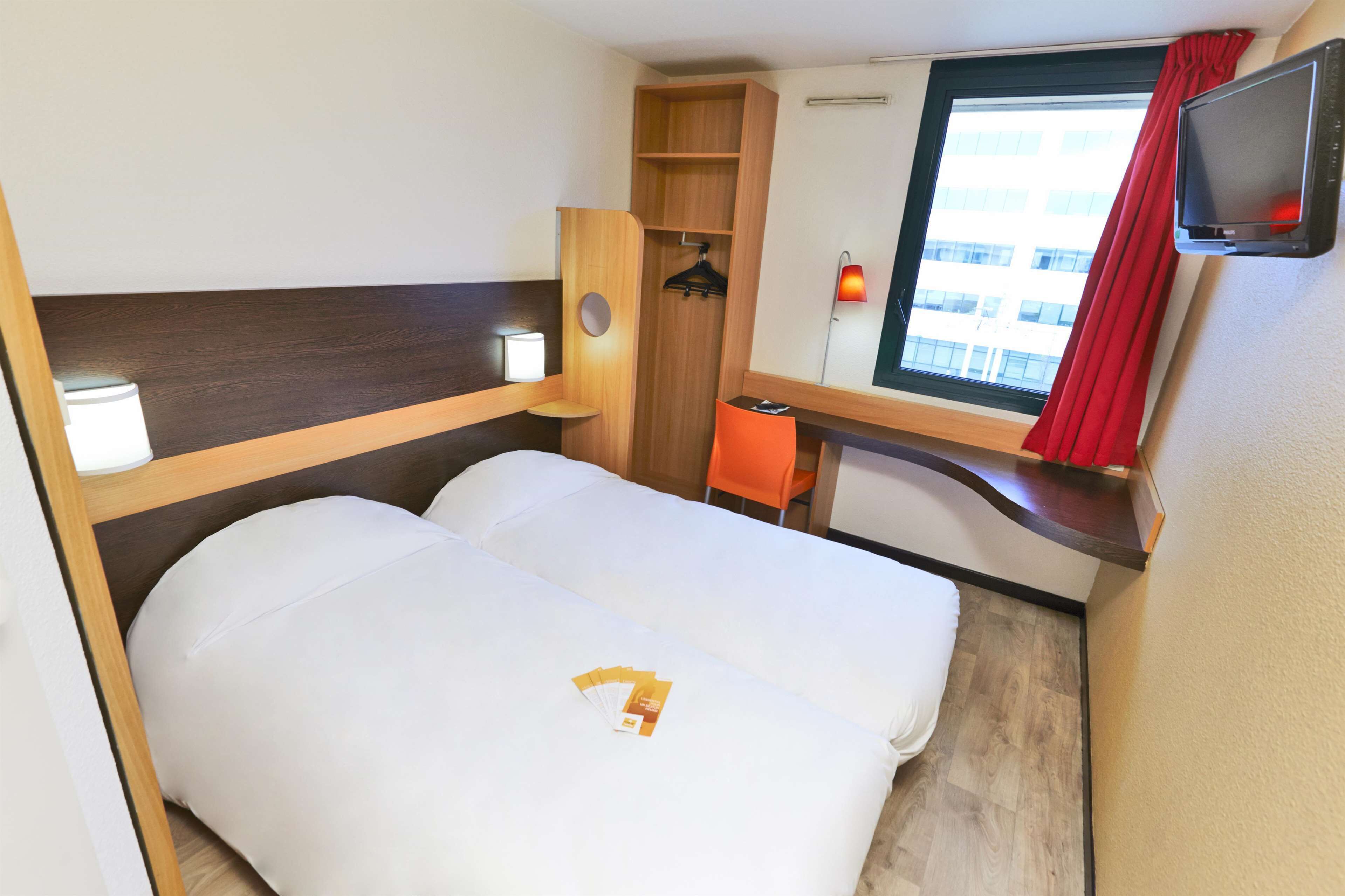 A compact hotel room with a bed, a small table, a chair, a wardrobe, and a TV mounted on the wall.