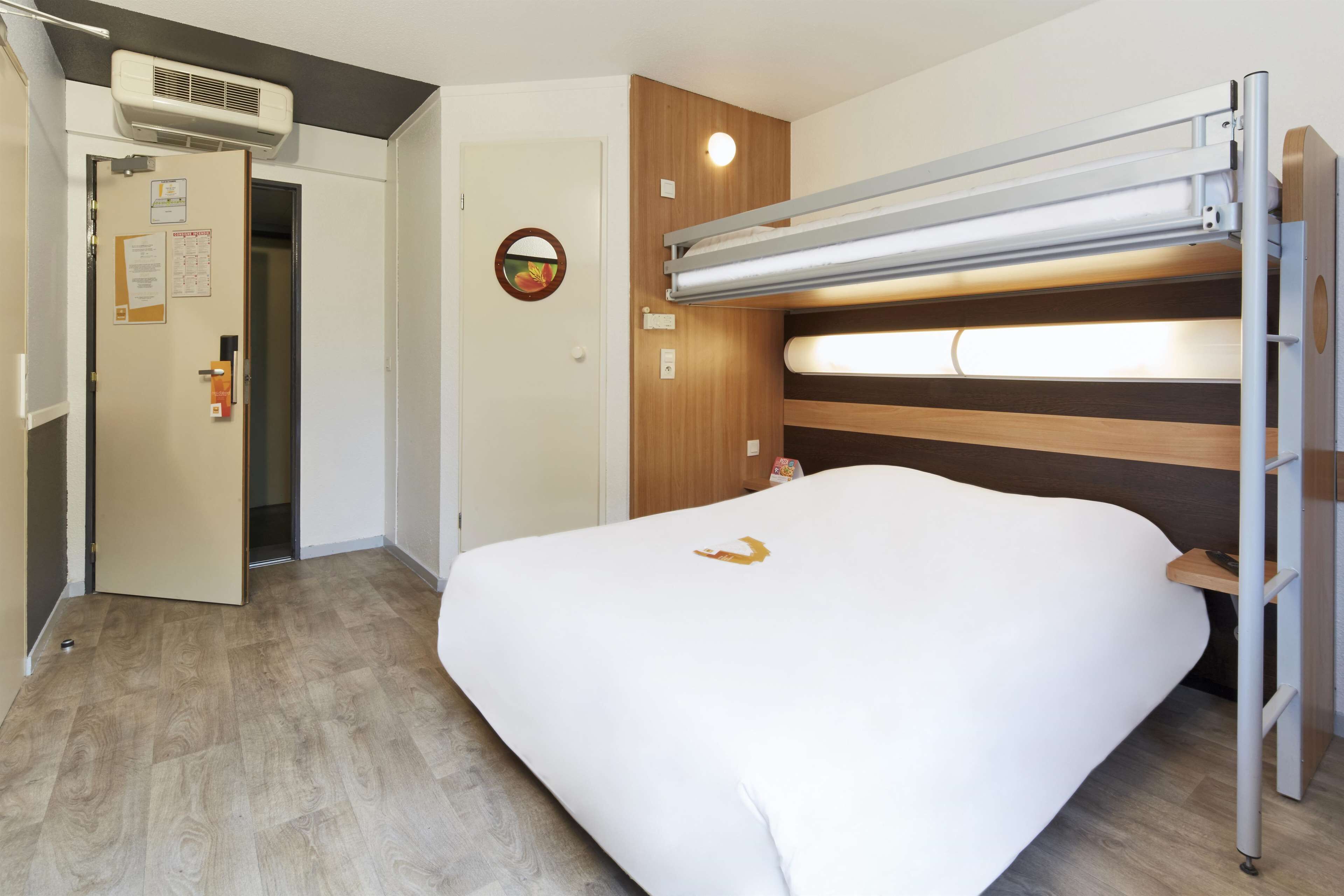 A hotel room with a bunk bed, a wooden headboard, a bedside table, and a door with a bulletin board.