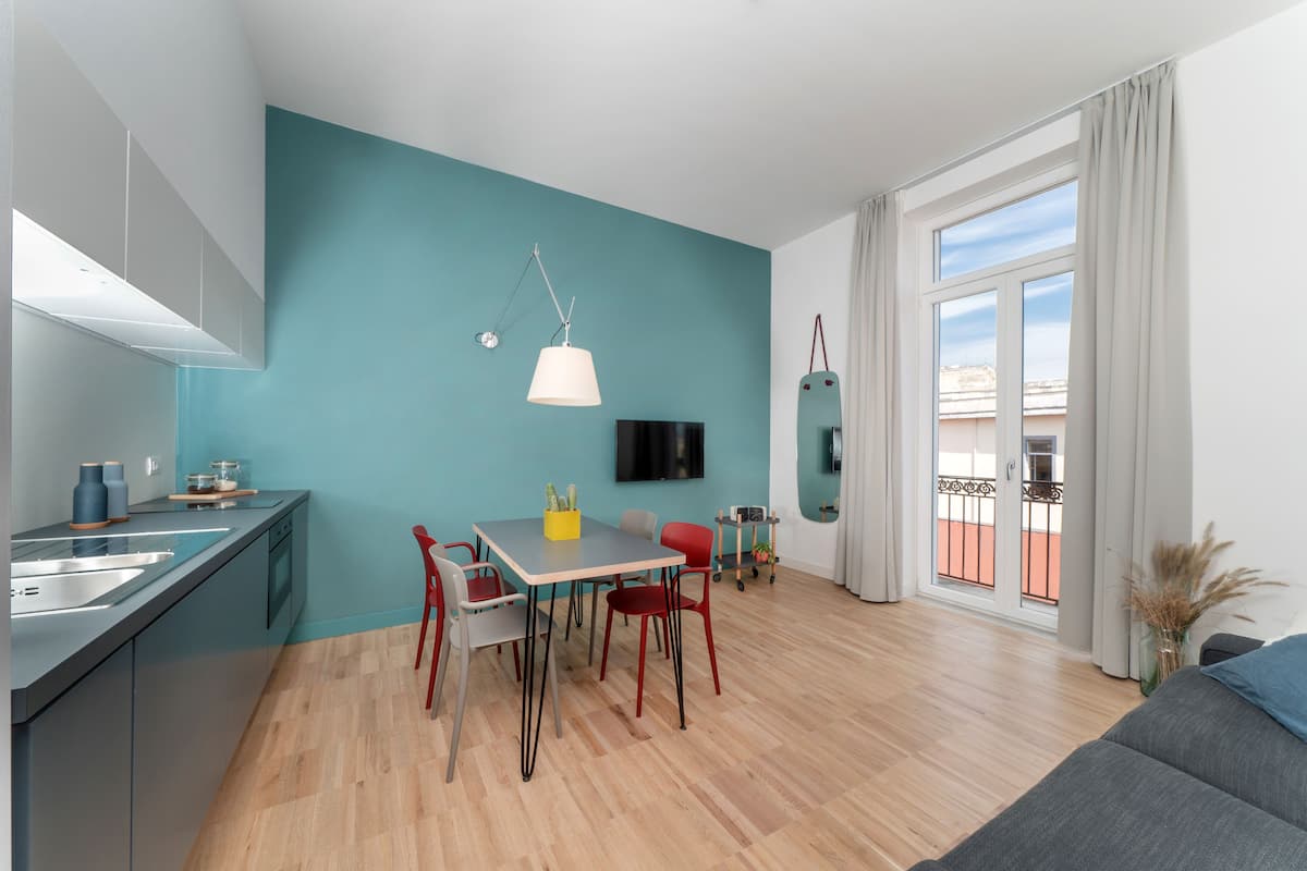 A modern kitchen with a teal accent wall, a dining area with a table and chairs, and a balcony with a view.